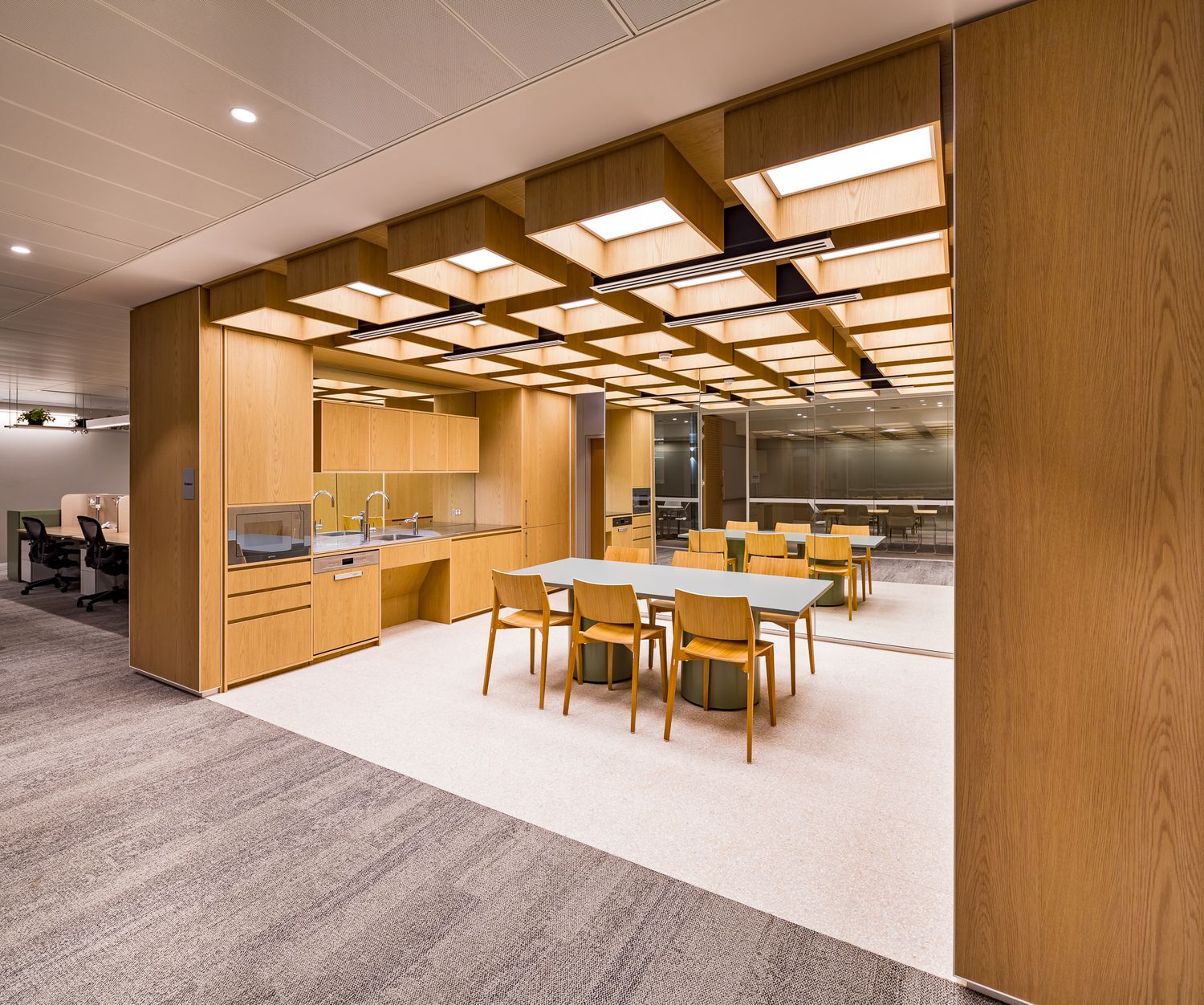 Open-plan office kitchen and meeting area with a timber grid ceiling, oak cabinets, wooden chairs and a central table.