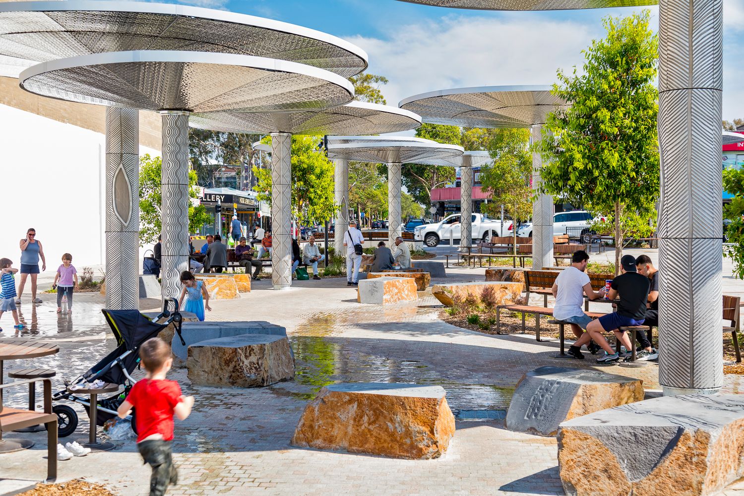 Families and children enjoy a shaded town square with mushroom-style canopies, boulder seating, benches and a stroller beneath green trees.