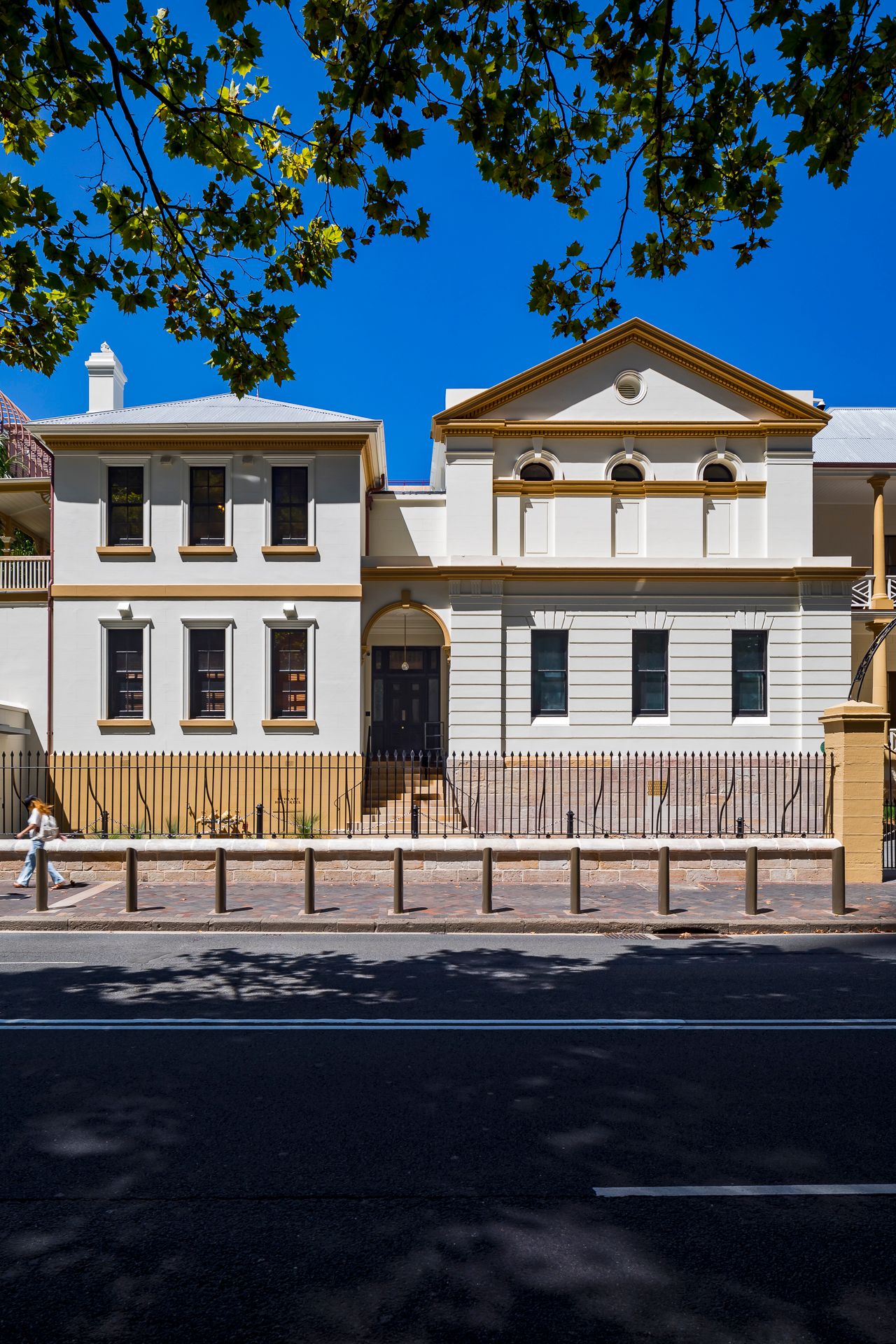 Two-story cream heritage building with yellow trim, arched entrance and pediment, behind an iron fence on a sunny tree-lined street.