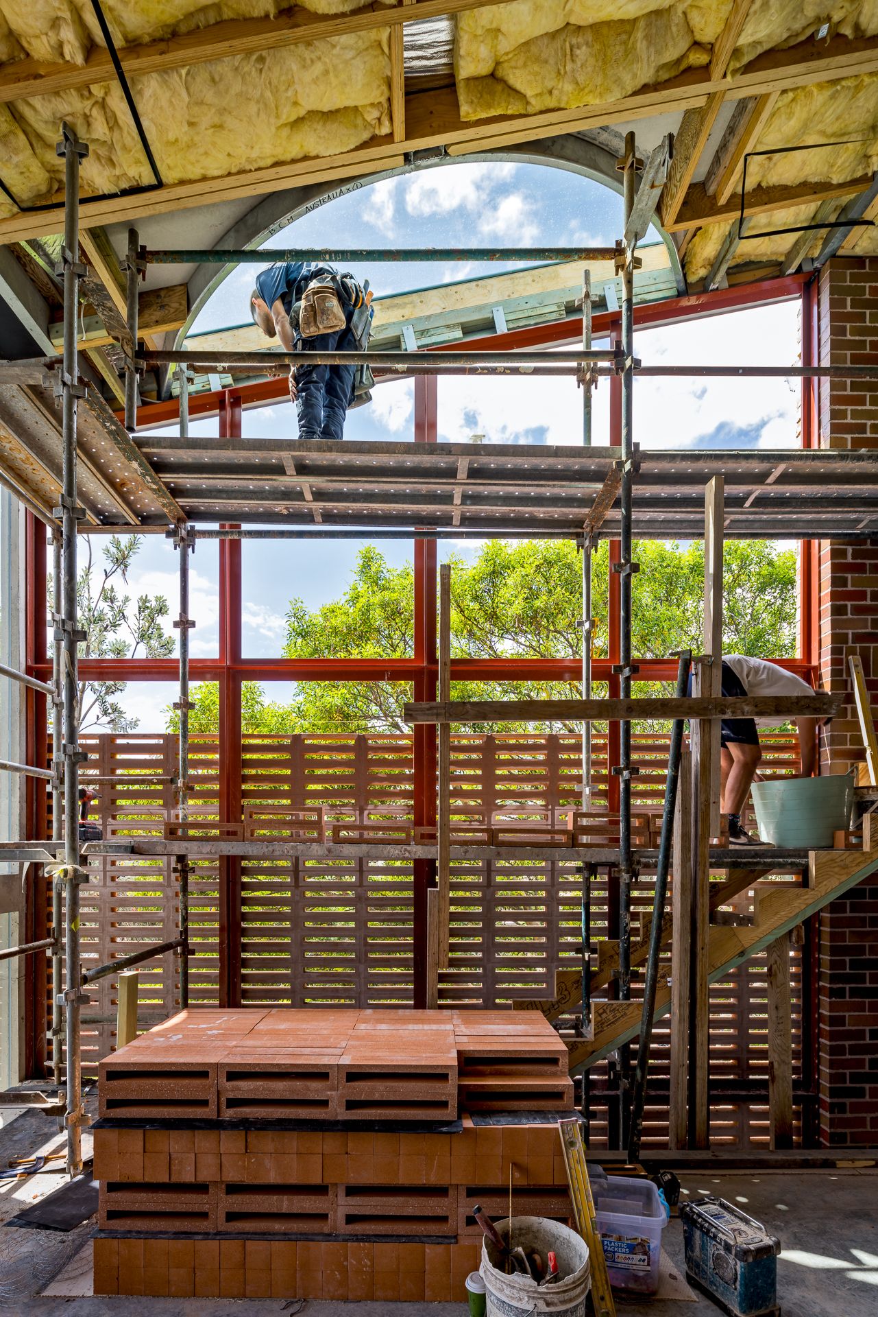 Construction workers on scaffolding inside a brick building with exposed insulation, arched window and timber slat screen under a blue sky.