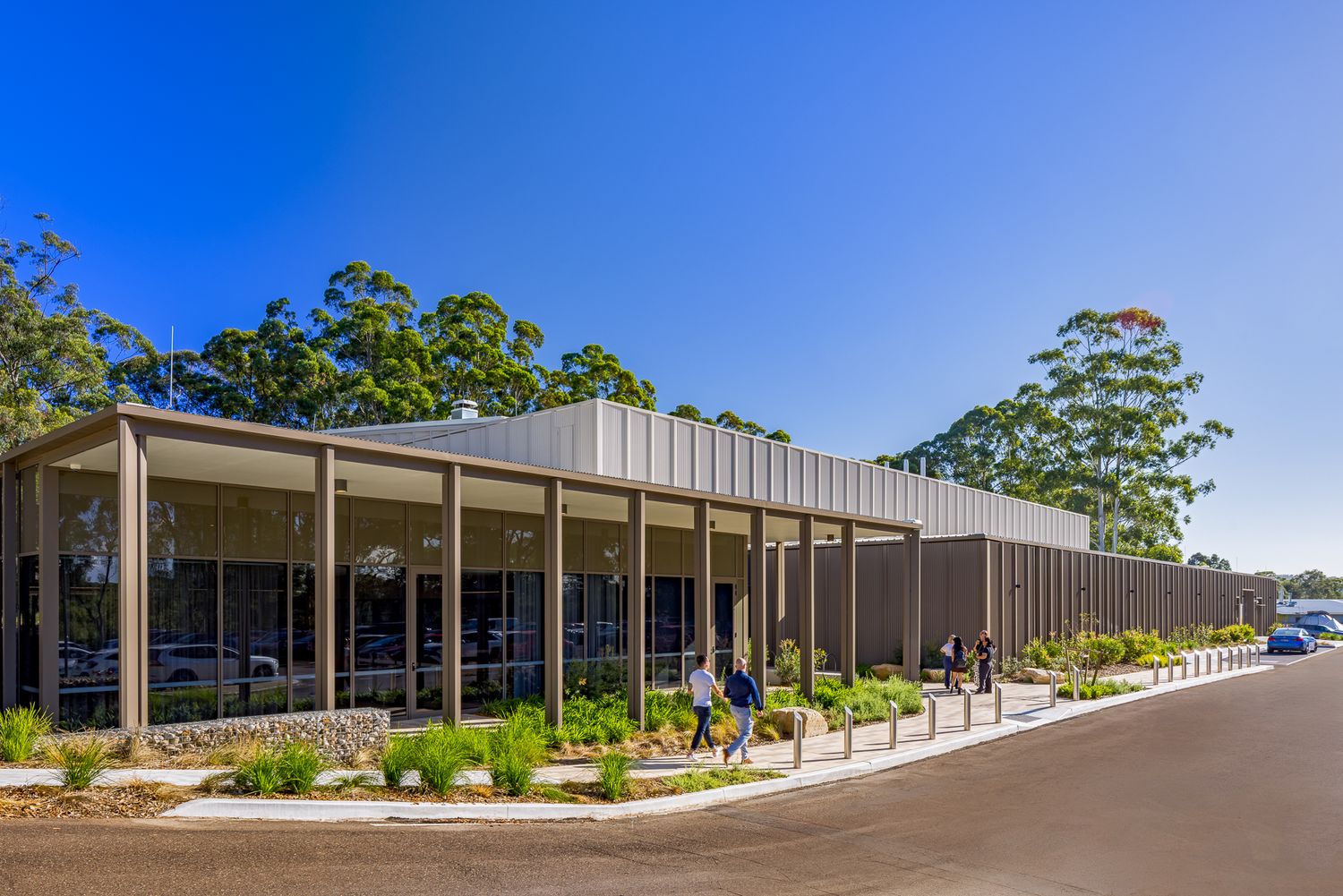 Visitors walk along a path beside a modern glass and timber-clad building framed by native gum trees and landscaped gardens.