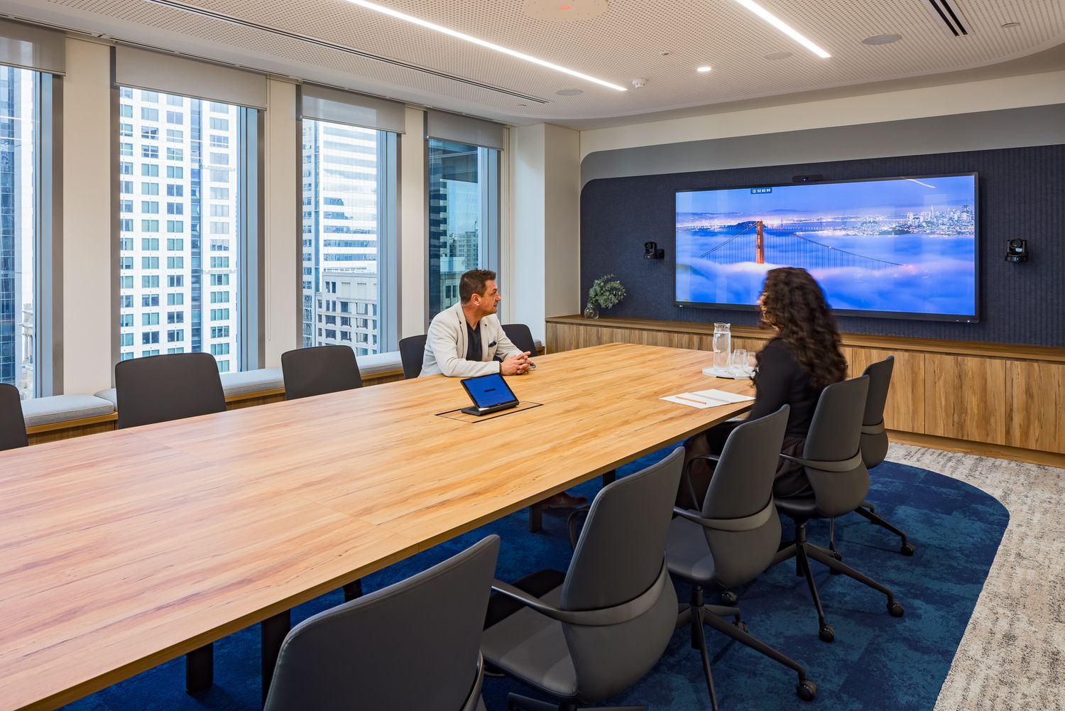 Two colleagues meet at a long wooden conference table facing a wall-mounted screen in a conference room with city skyline views.