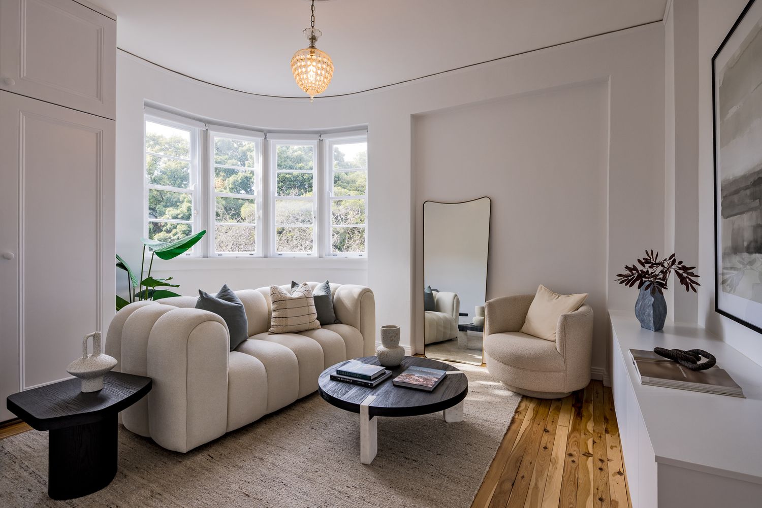 Elegant sitting room featuring a curved cream sofa, bay windows, a tall floor mirror, a swivel armchair and a dark side table.