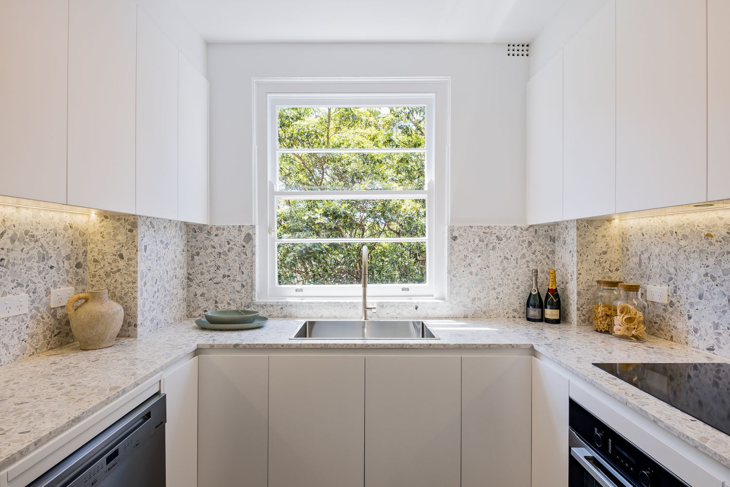 Modern white kitchen with stone benchtops, speckled splashback, undermount sink and window overlooking green trees.
