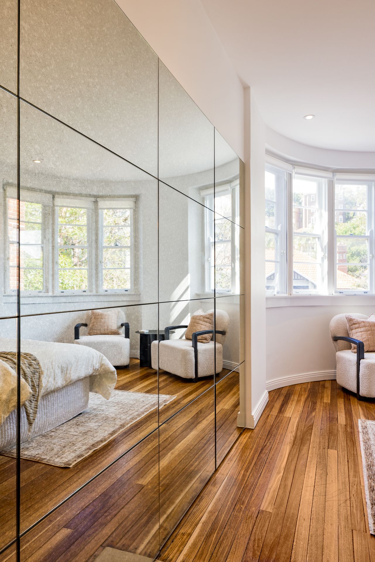 Mirrored wall panels reflect a bedroom bay window and armchairs beside a curved wall and polished timber floorboards.
