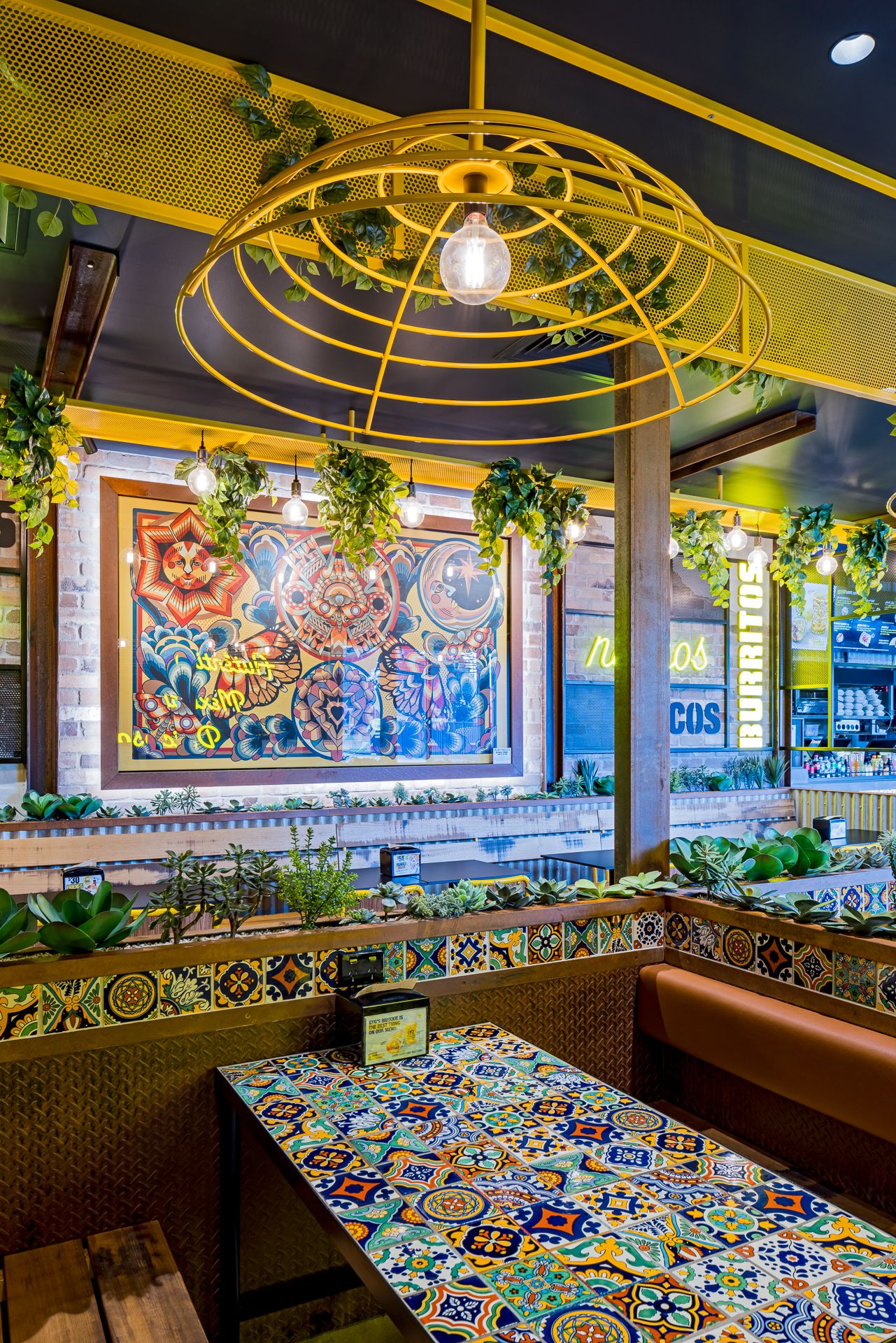 Mexican restaurant dining area with a bold painted mural, talavera-style tiled table and a yellow wire pendant light.