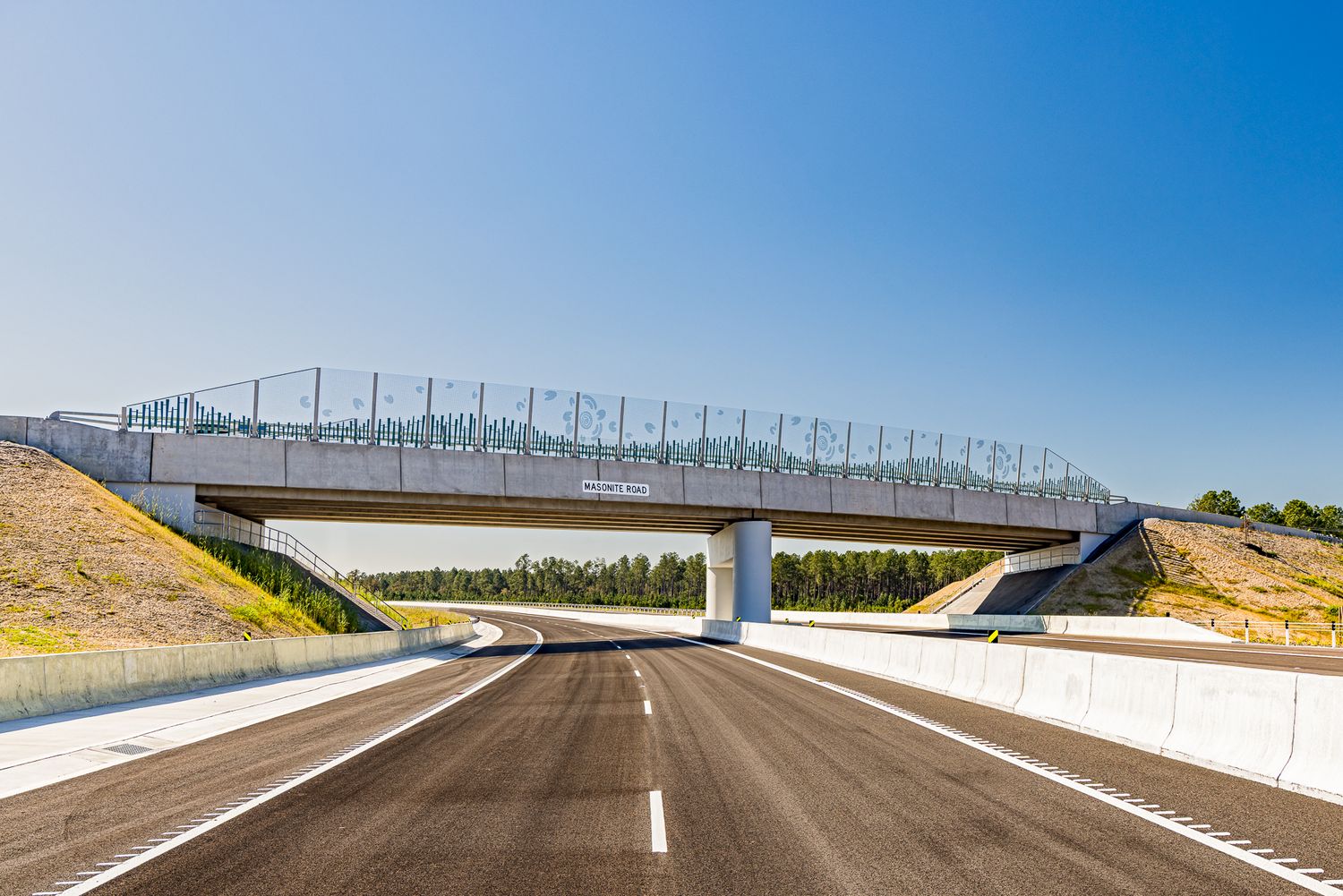Newly built asphalt highway passing beneath a concrete overpass with safety fencing, set between grassy embankments under a clear blue sky.