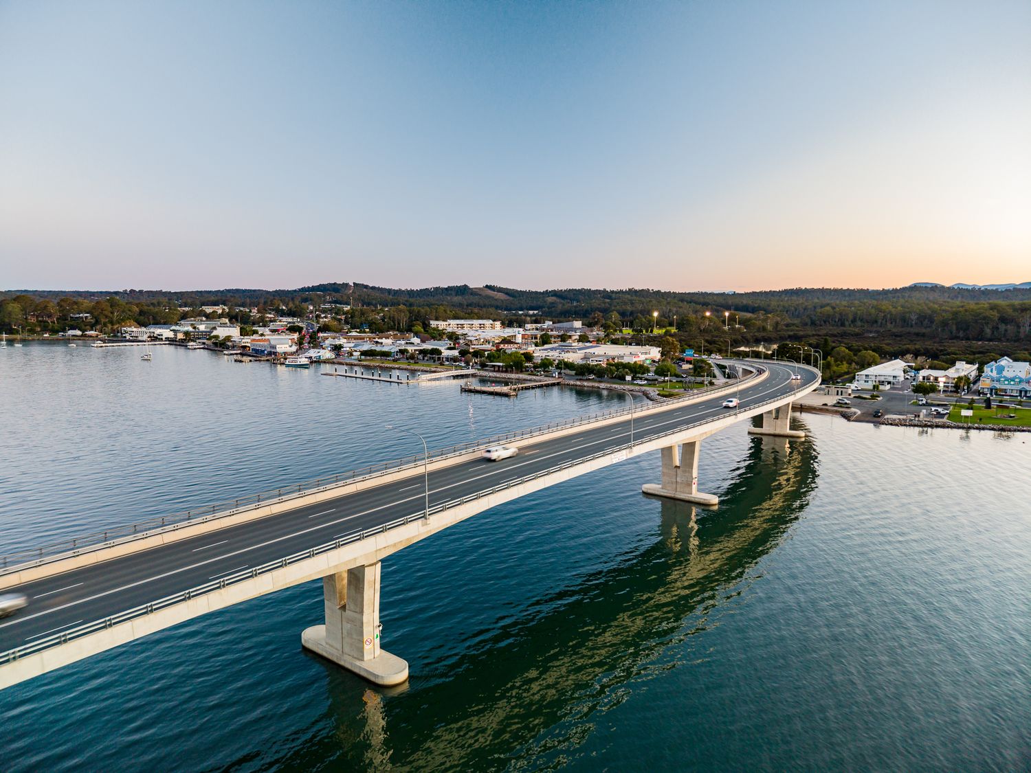 Aerial view of a curved concrete bridge over calm water leading to a coastal town at sunset.