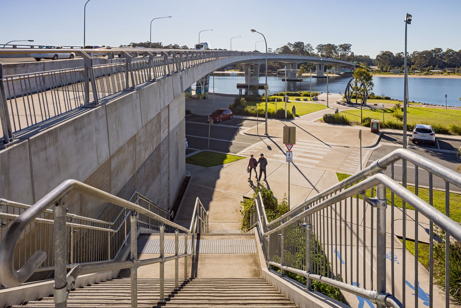 Two people walk along a path beside a curved concrete bridge with stairs, railings and a calm waterway beyond.