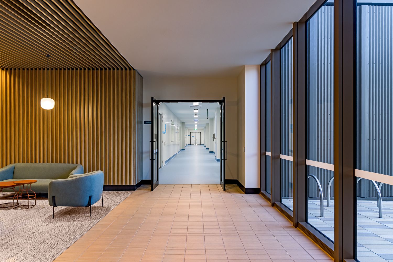 Timber-slatted corridor with a lounge seating area and floor-to-ceiling windows leading to an open doorway and a long hallway beyond.
