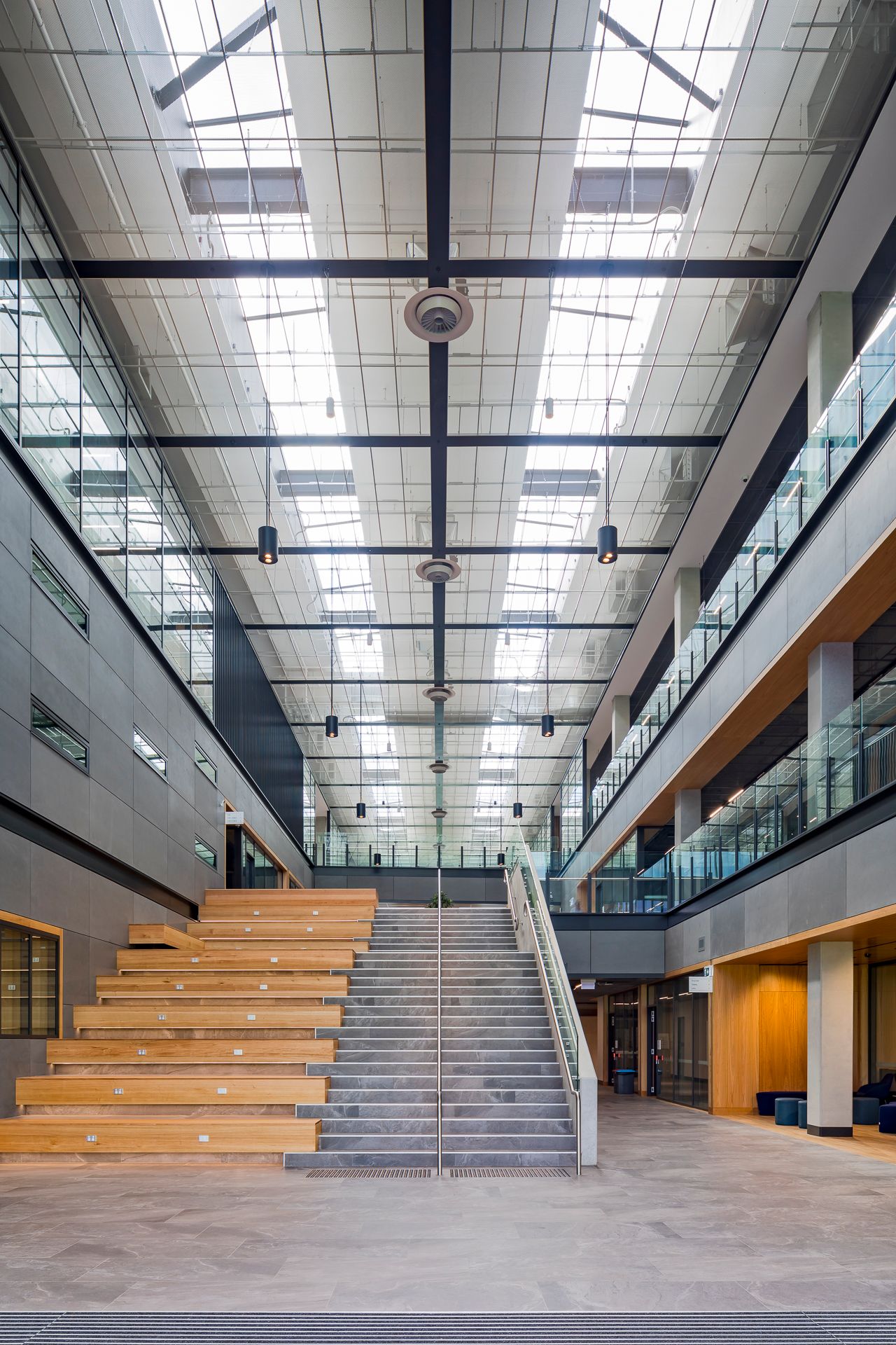 Tall skylit atrium in a modern educational building with a staircase, timber seating steps, and glass-fronted balconies on both sides.