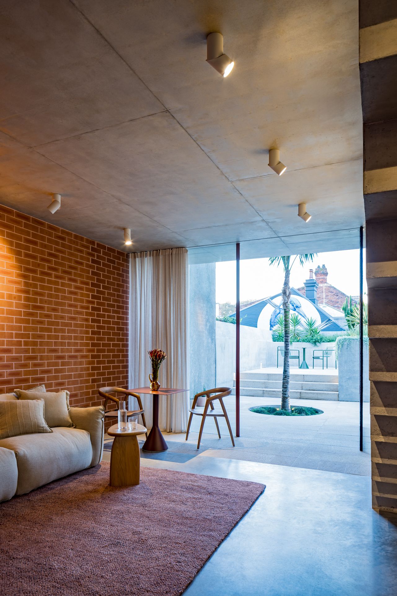 Open-plan living area with concrete ceiling, exposed brick wall, cream sofa and floor-to-ceiling glass doors opening to a sunny courtyard.