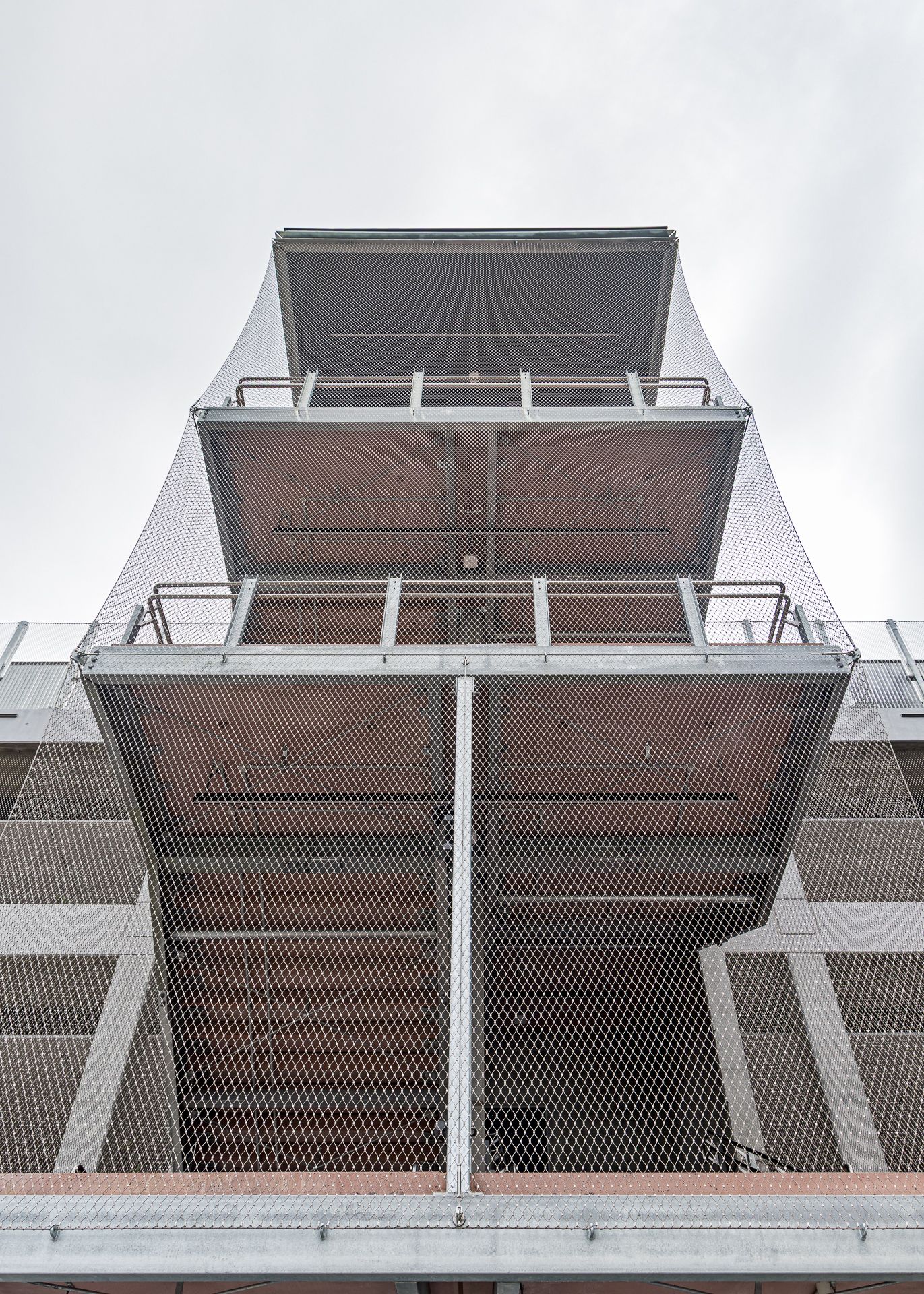 Looking up at a stair tower with layered landings, galvanized railings, and mesh screening against an overcast sky.