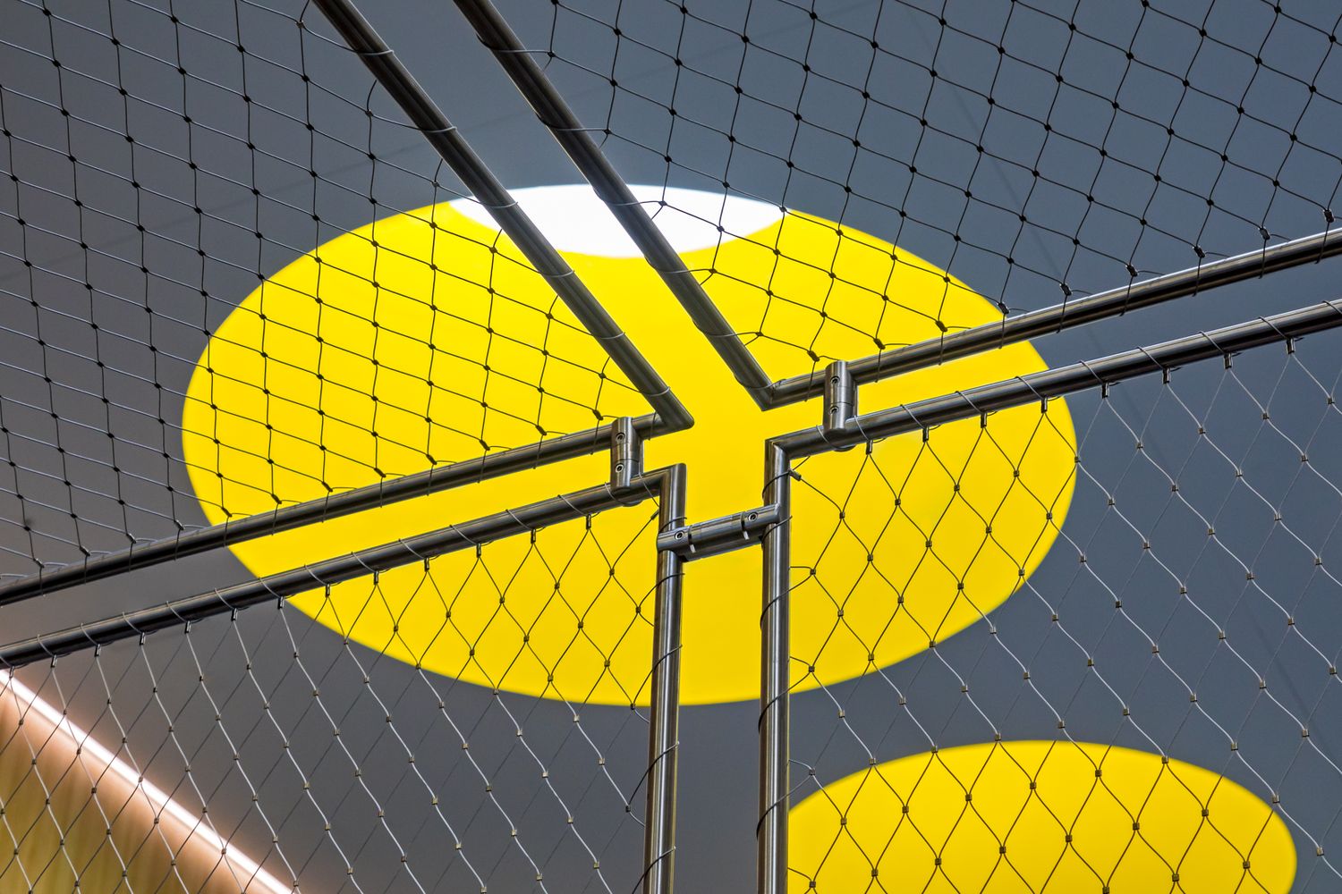 Looking up through steel mesh netting at bold yellow circular shapes against a dark gray ceiling in a modern architectural space.