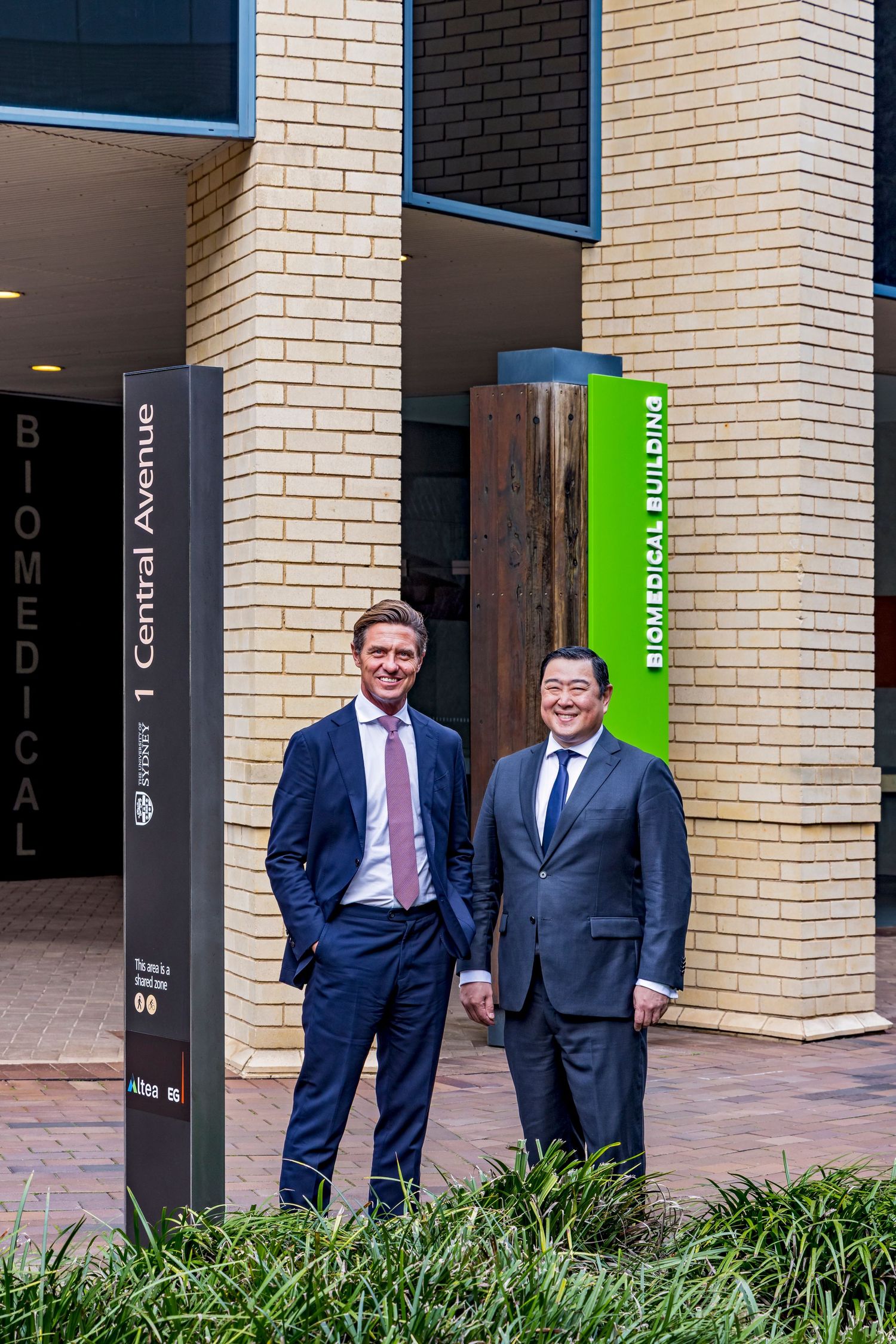 Two men in business suits stand smiling outside the Biomedical Building entrance next to a 1 Central Avenue directory sign.