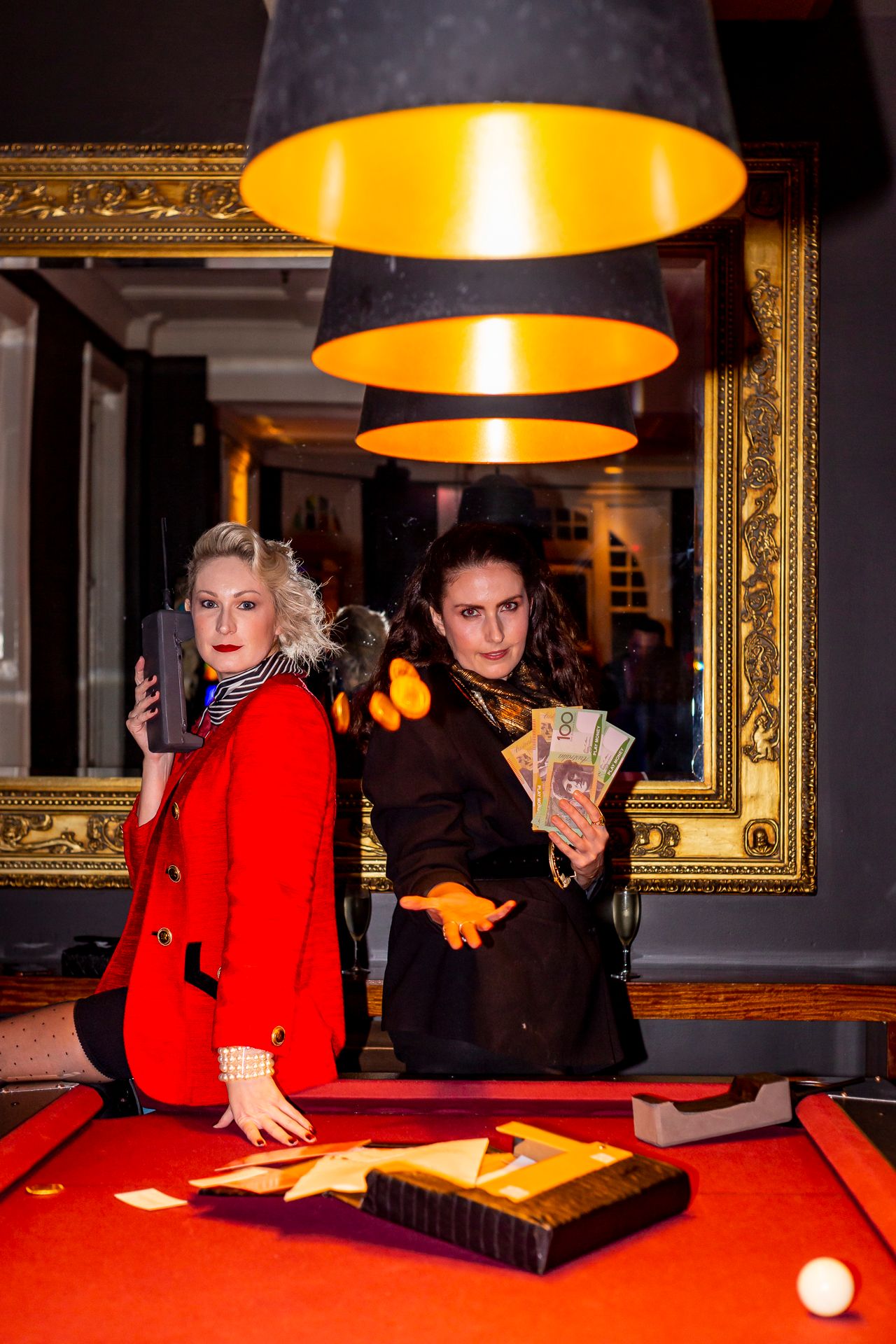 Two women in stylish coats pose at a red pool table, one holding a phone and the other fanning out cash beneath glowing pendant lamps.