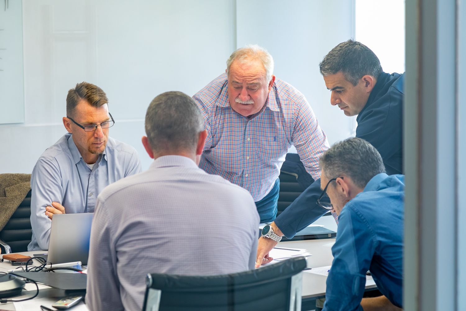 Five men in business casual clothing huddle around a table in a meeting room, discussing work together near a laptop.