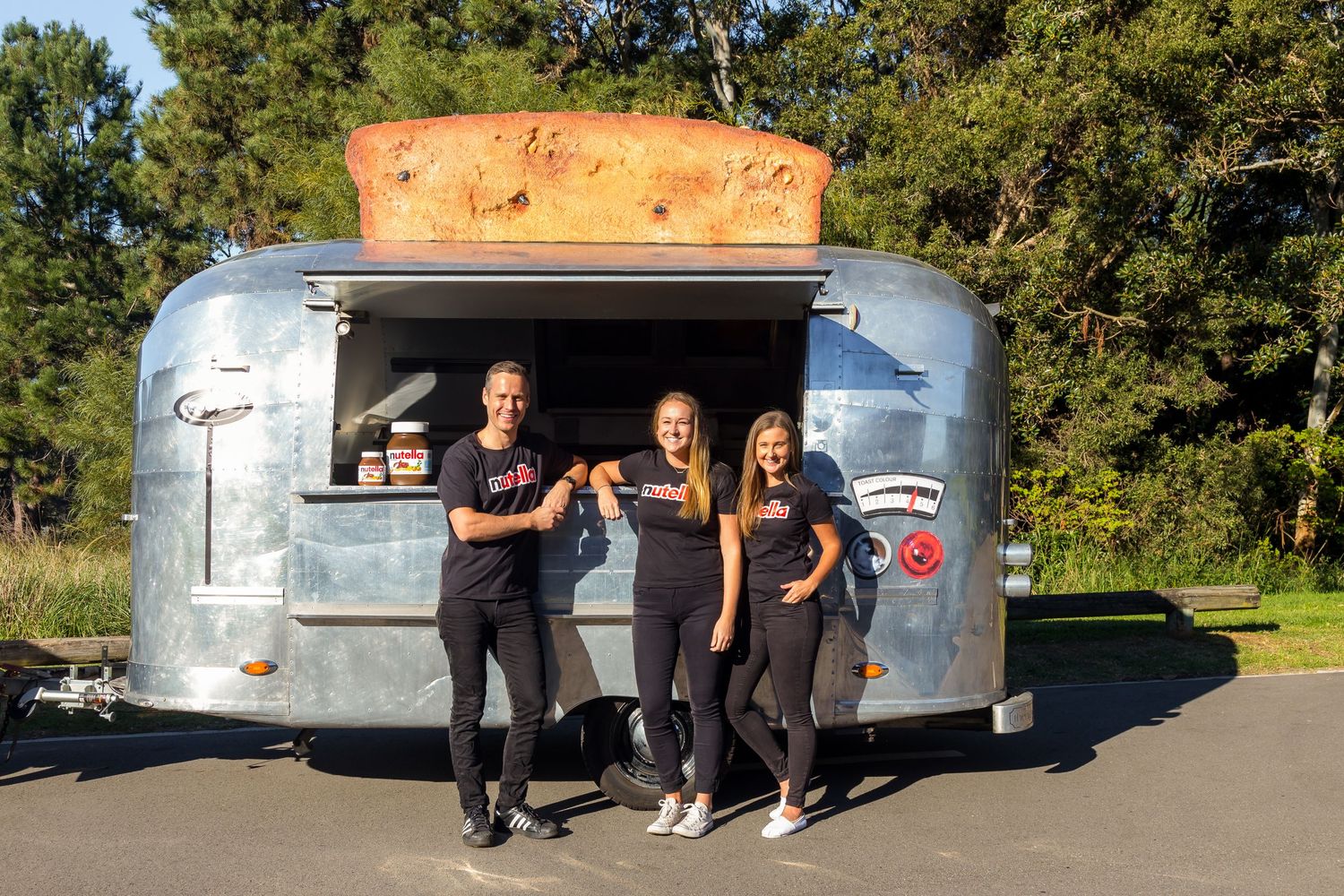 Three smiling staff members stand in front of a vintage silver Airstream food trailer topped with a giant bread loaf.