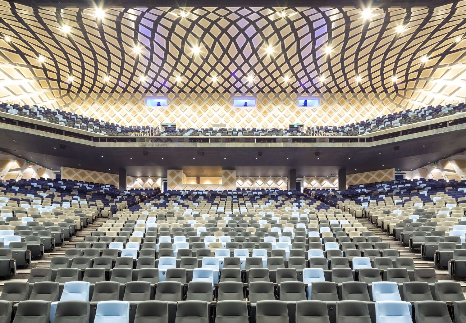 Large tiered convention theatre with a sculptural lattice ceiling, rising seating and screens, viewed from the stage.