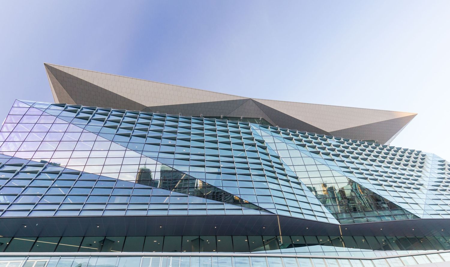 Low-angle view of a modern glass building with a faceted facade and angular metallic roof under a blue sky.