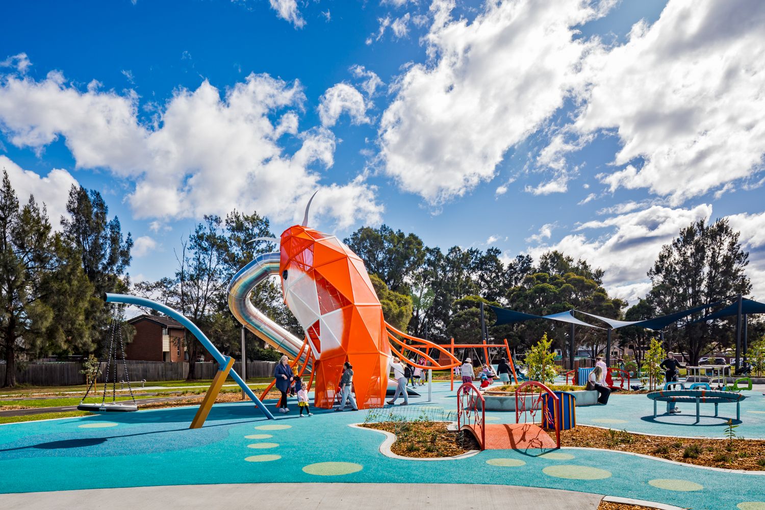 Colorful children's playground with a giant orange fish-shaped climbing structure, slide and rubber surfacing under a blue sky.