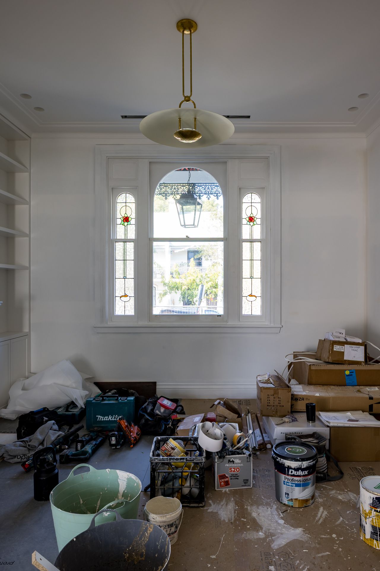 Renovation work in progress in a room with stained glass windows, a brass pendant light, paint cans, tools and boxes on the floor.