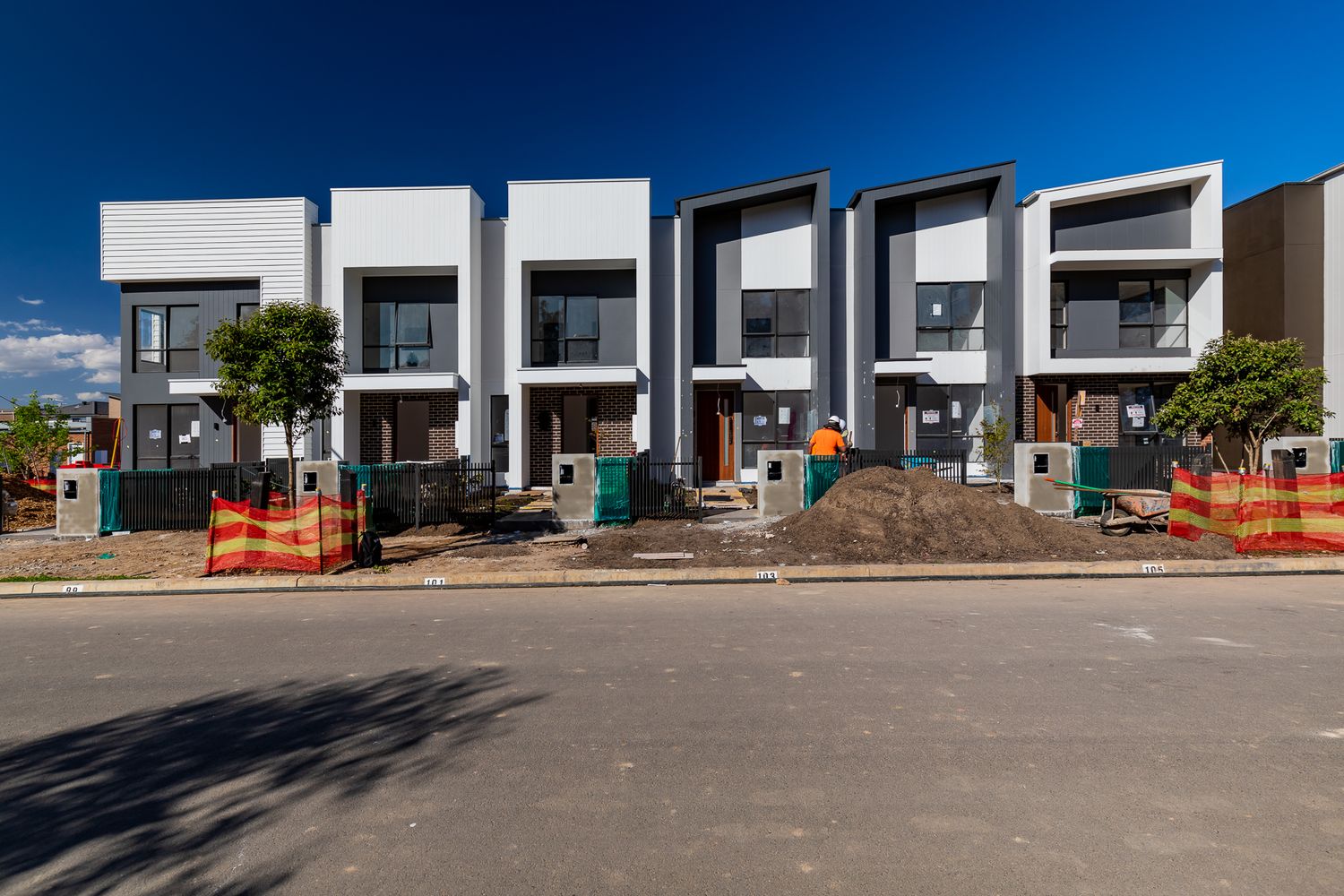 Row of modern white and dark gray townhouses under construction with orange safety fencing and mounds of soil out front.