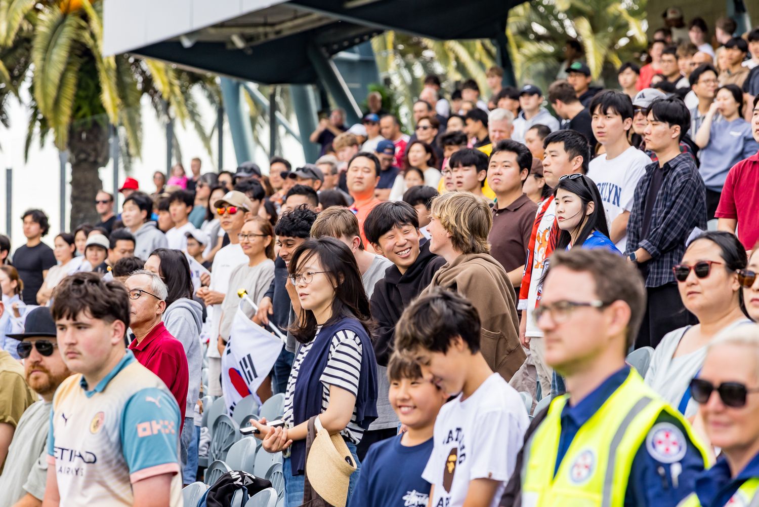 Crowd of football fans watch a match from the stadium stands while a security officer in a high-visibility jacket stands nearby.