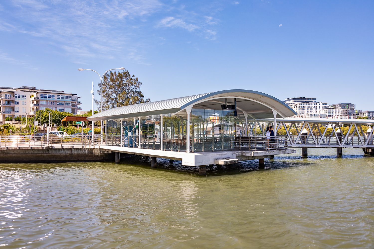 Riverside ferry terminal with a curved metal roof and glass walls on a timber pier, with apartment buildings and trees behind.