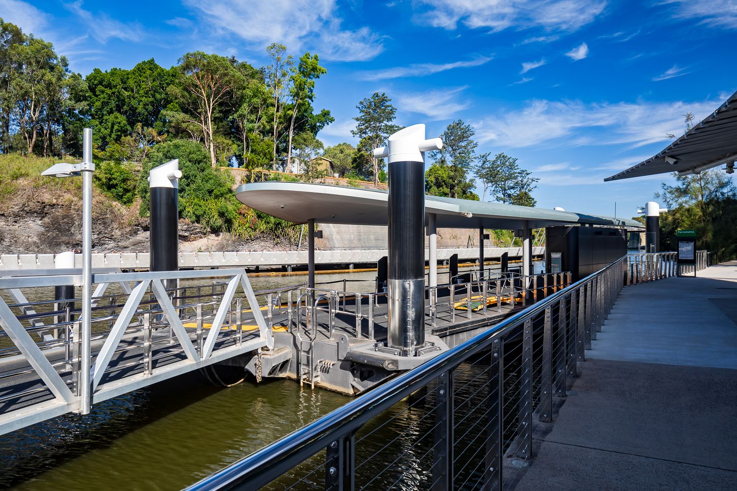 Ferry wharf pontoon with white-capped black piles and a gangway beside a leafy riverbank and calm green water.
