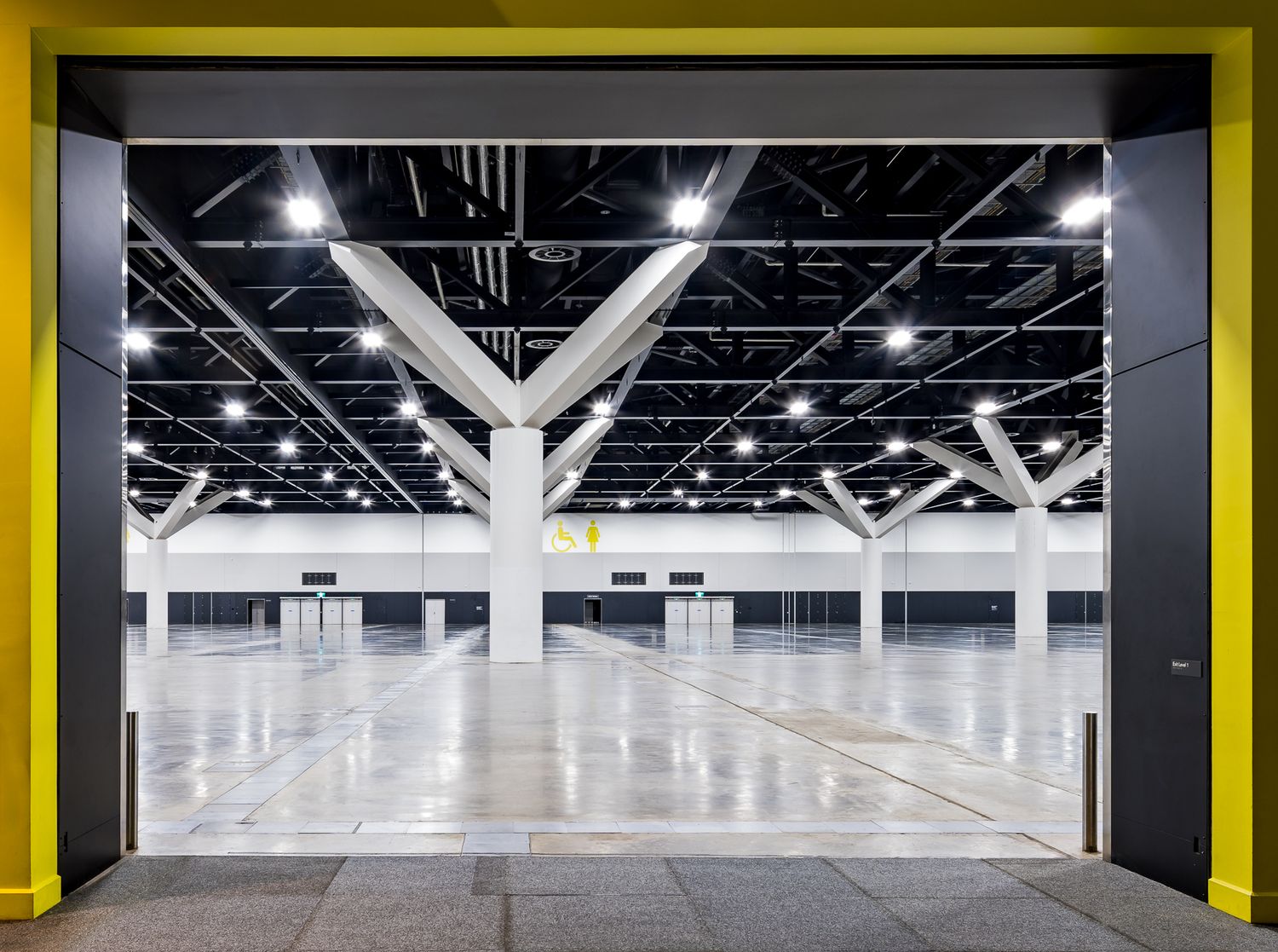 Framed view into a vast empty exhibition hall with a polished floor, white tree-like support columns and bright ceiling lights.