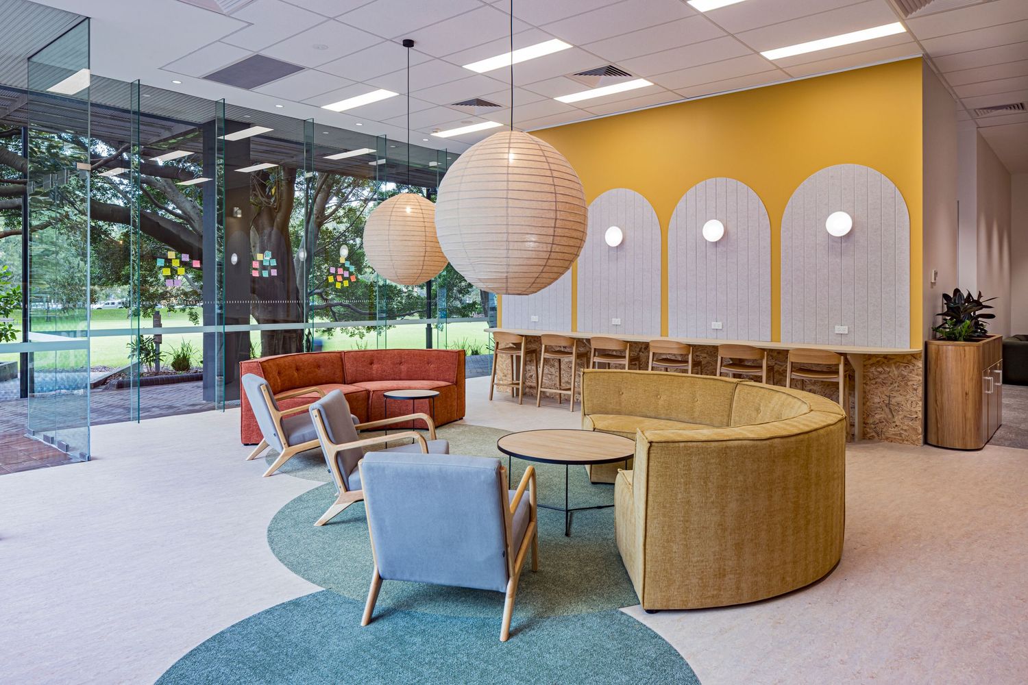 Bright office lounge with curved tan banquette seating, orange sofa, pendant lamps and large windows overlooking trees.