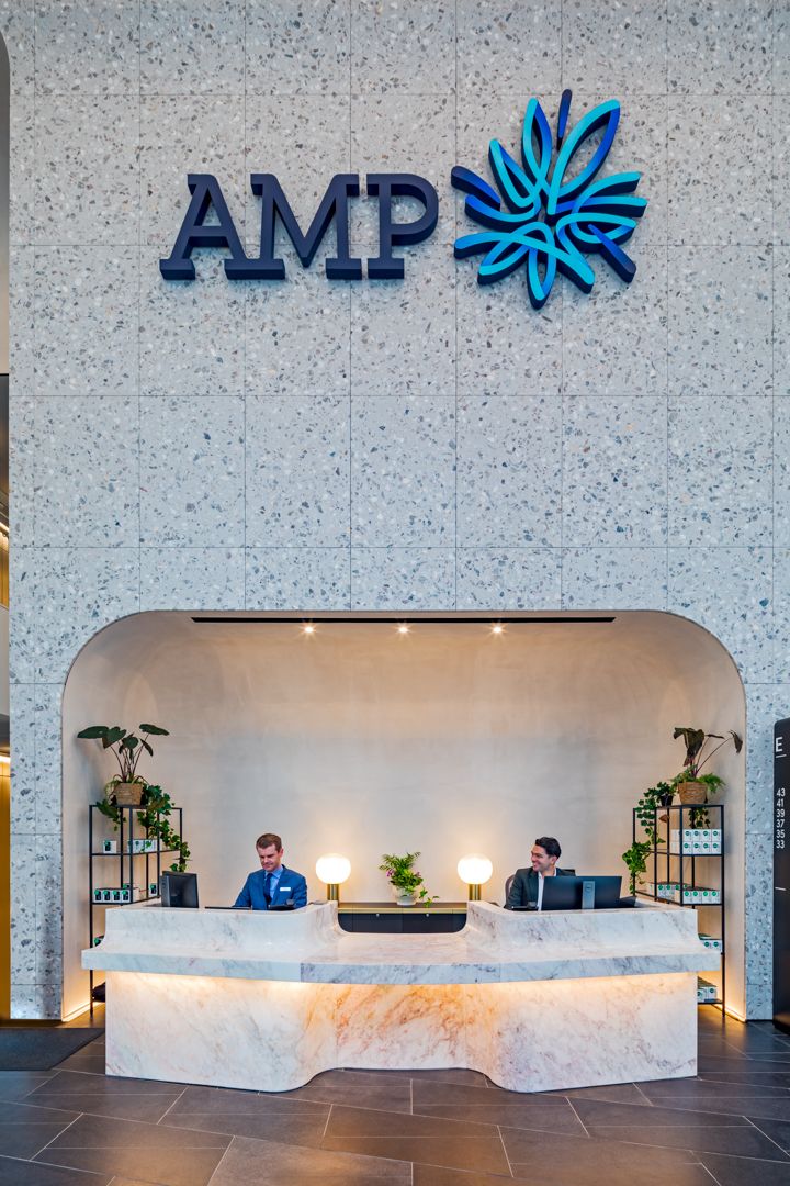 Two receptionists work at a marble reception desk below the illuminated AMP logo on a terrazzo wall in a modern corporate lobby with plants.