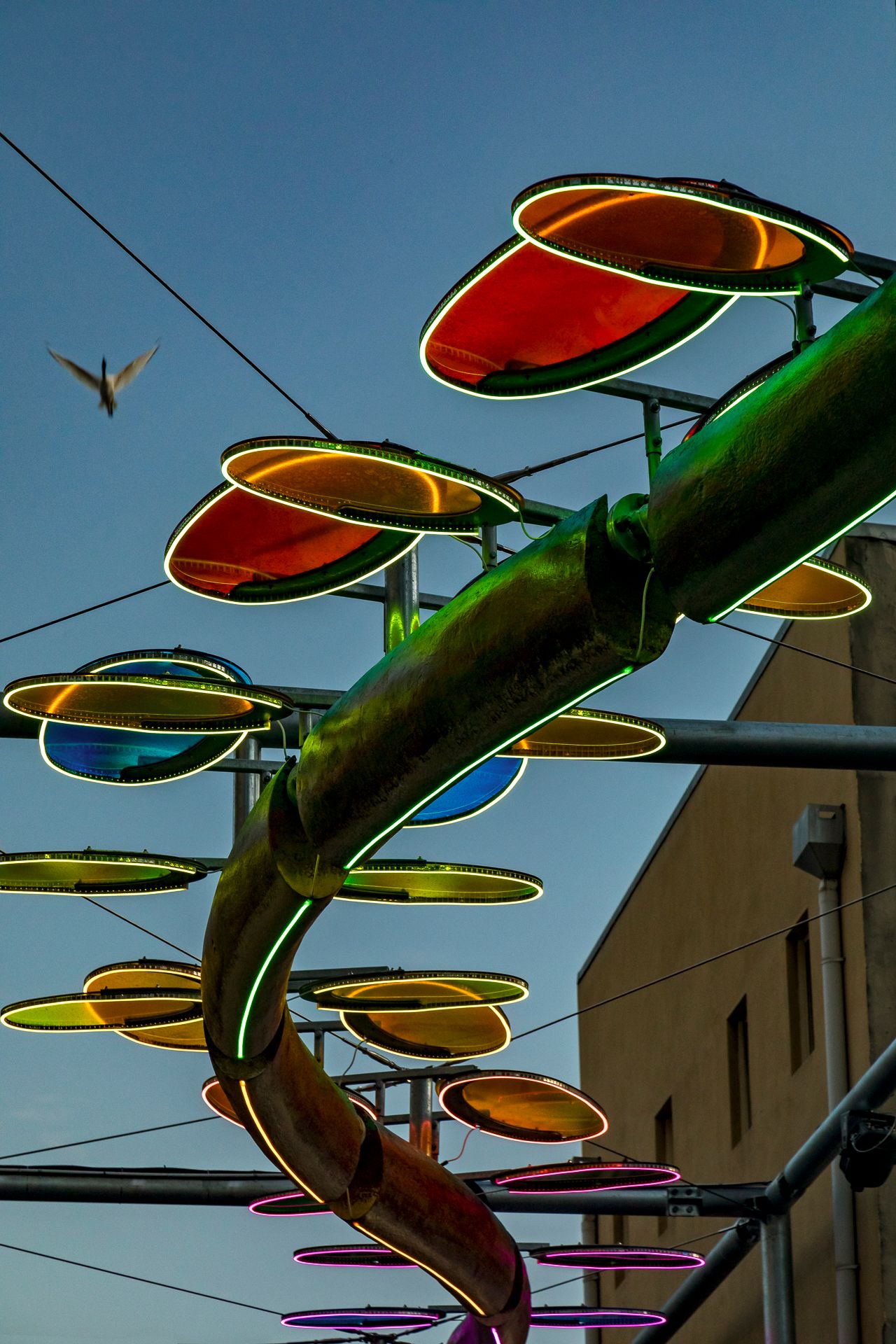 Close-up of a glowing serpent sculpture with green-rimmed red and orange oval discs against a dusk sky with a bird flying.