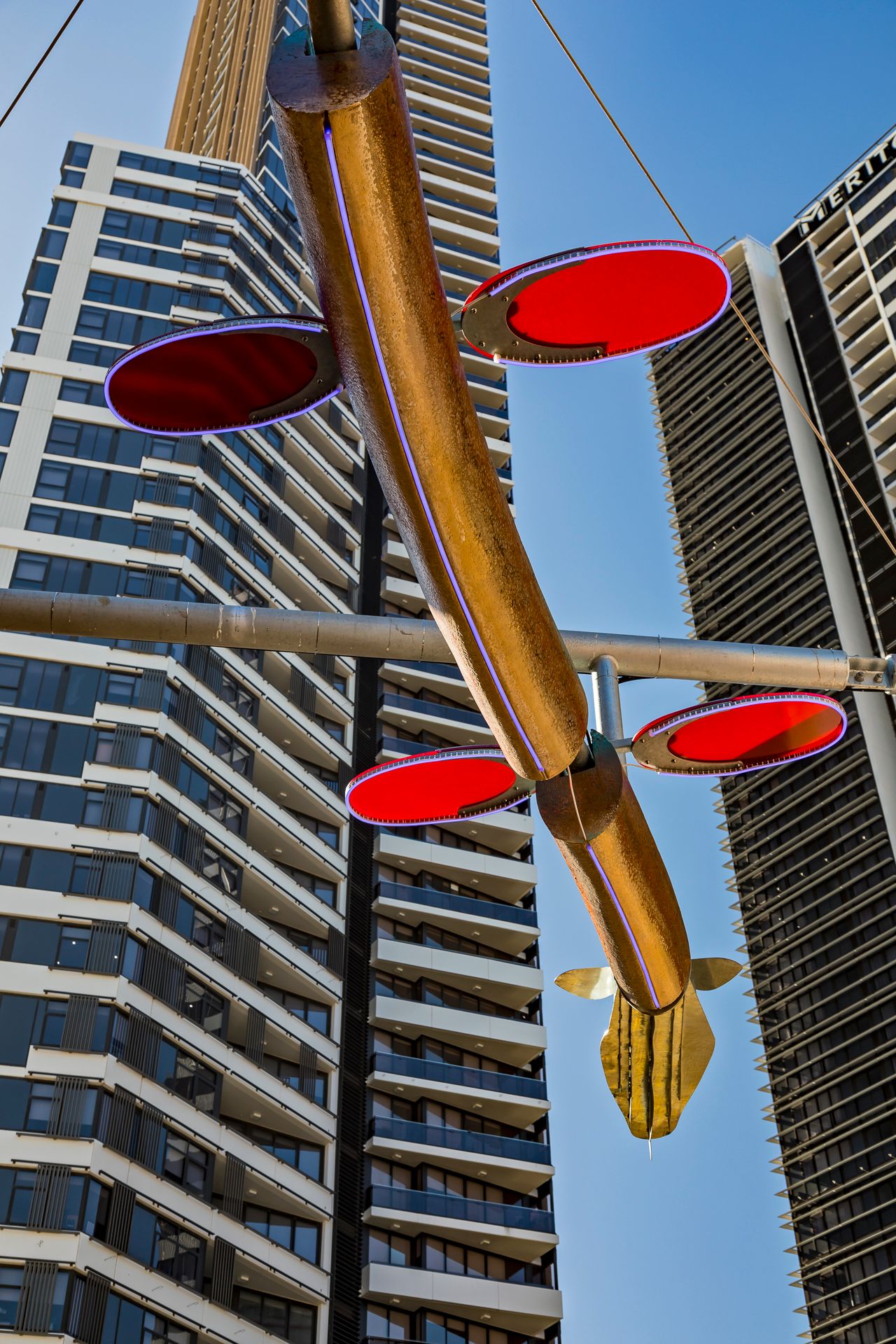 Red glowing discs and a golden fish-shaped tail on a hanging sculpture framed by tall apartment towers.
