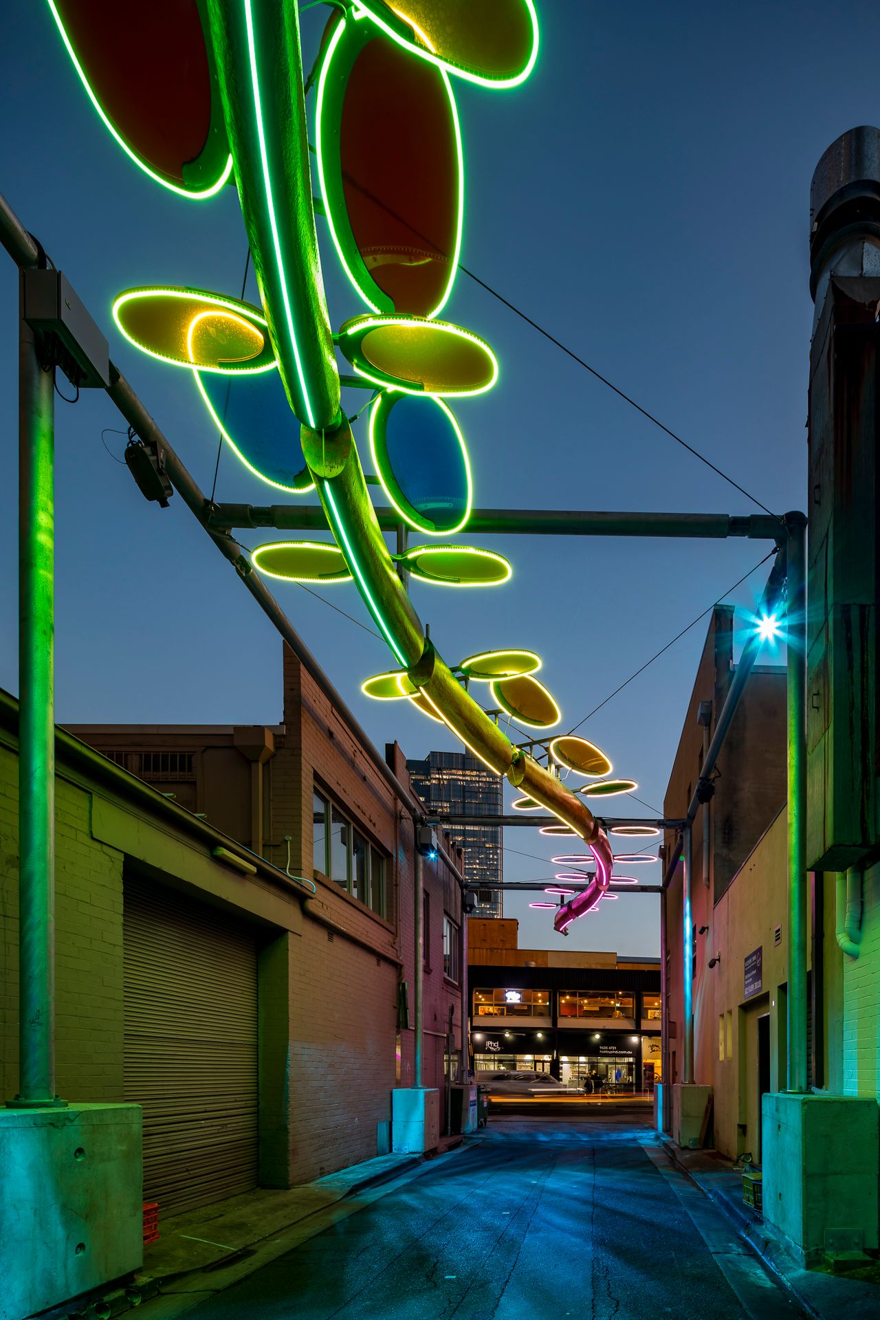 Green glowing serpent-shaped light sculpture with colorful oval discs hanging above a city laneway at twilight.