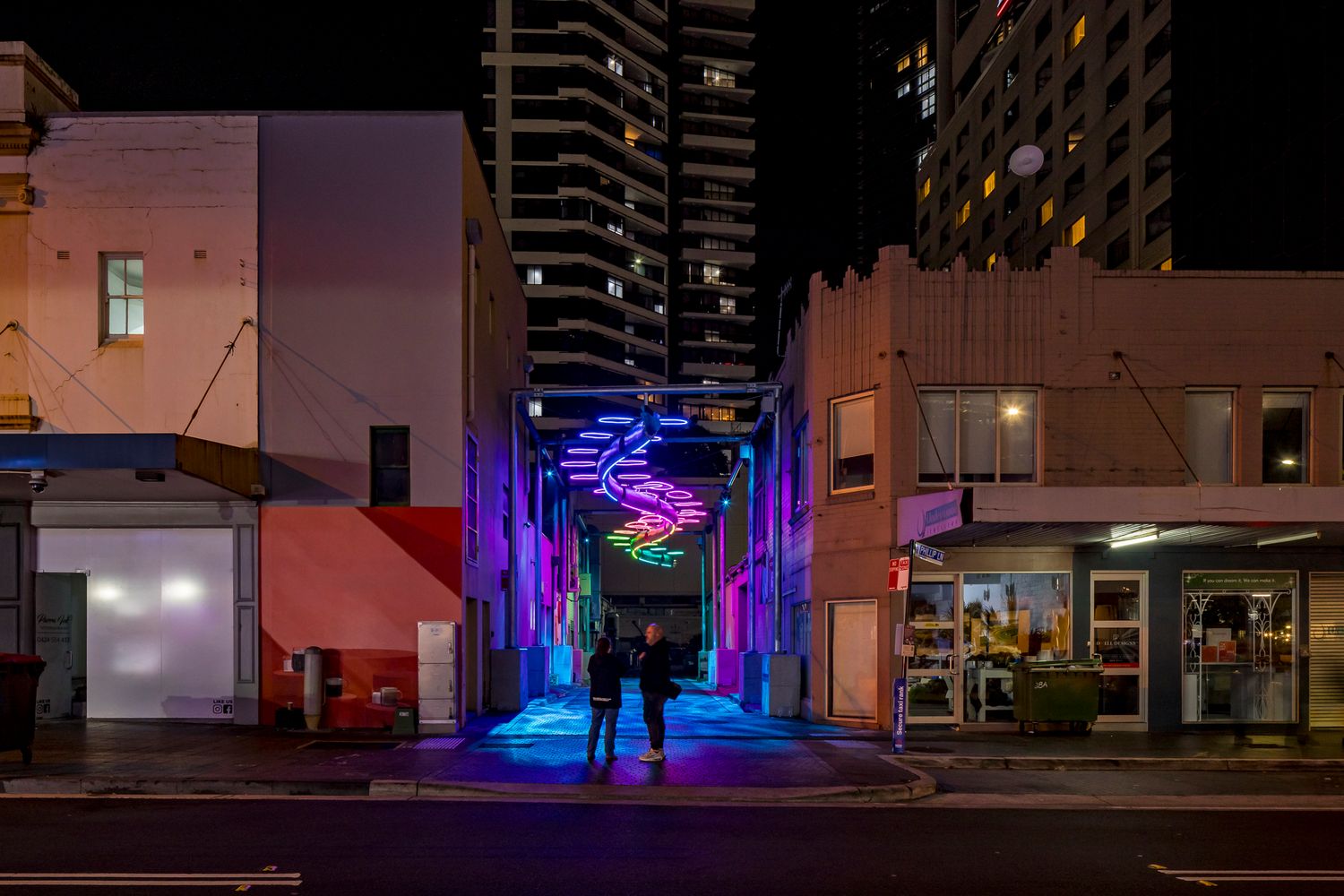 Two people walk toward a purple and blue neon light artwork hanging over an urban laneway beneath tall city towers at night.