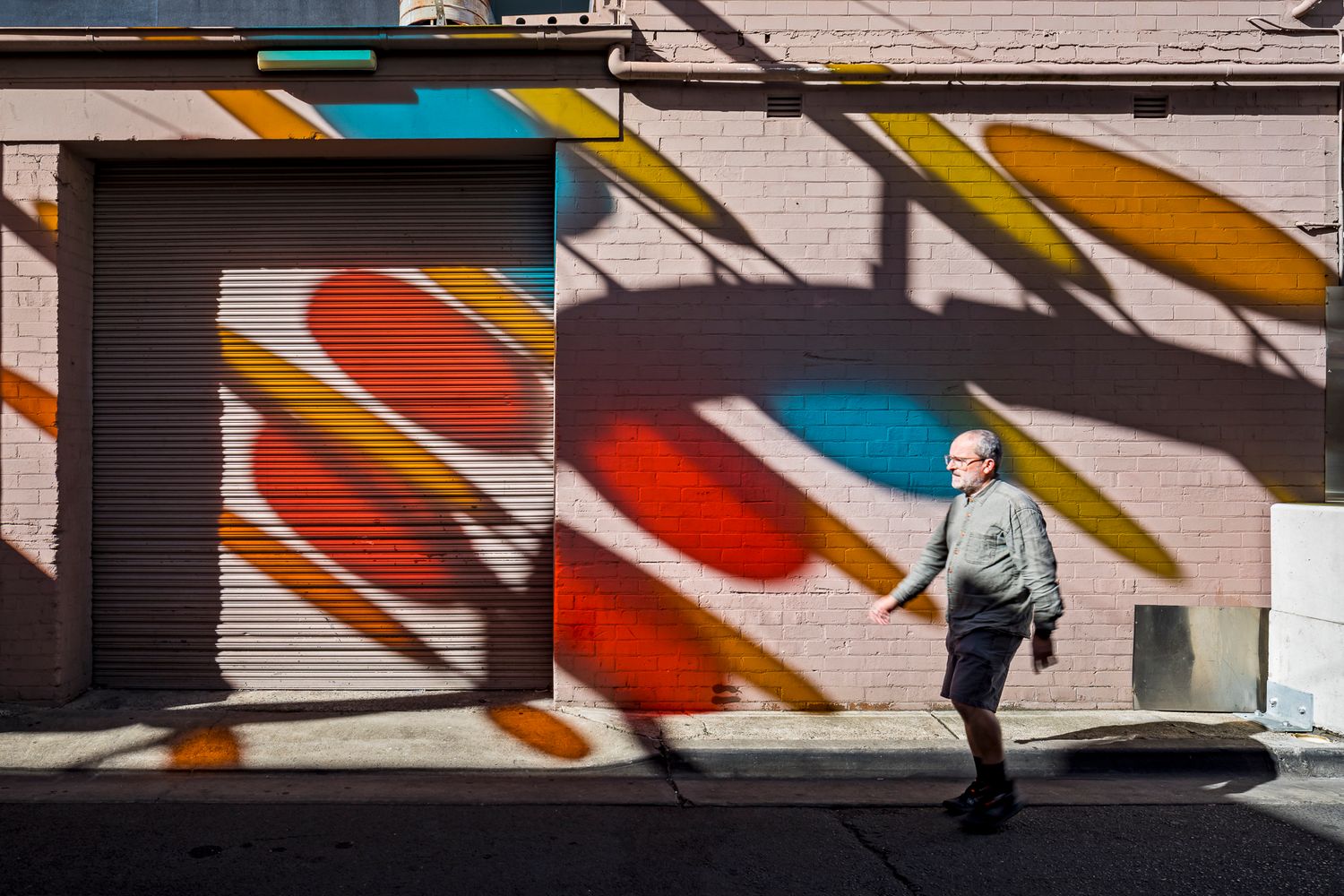Man in a gray sweater walks past a brick wall and roller shutter covered in colorful oval shadows and reflections.