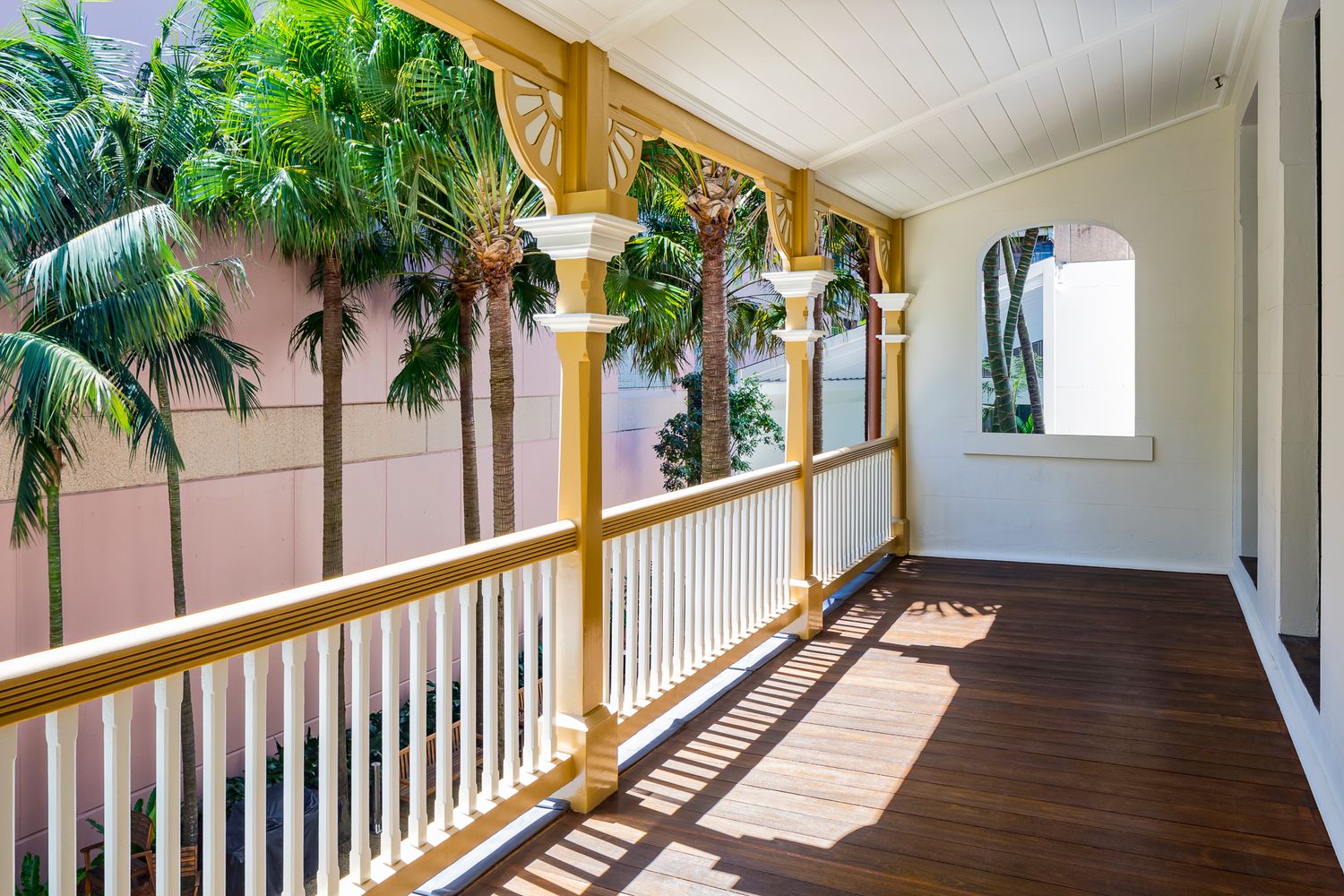 Sunlit timber verandah with white balustrade, golden ornamental posts and a polished dark wood floor overlooking tall palm trees.