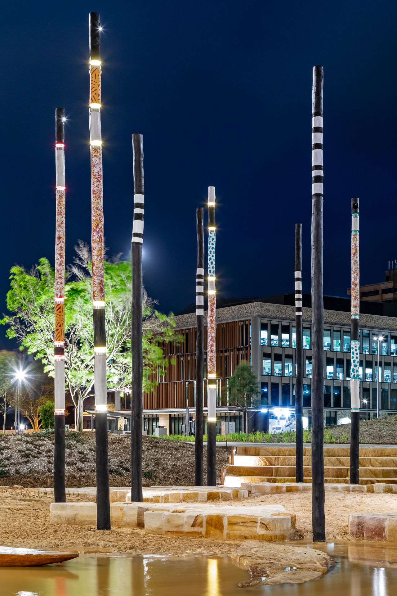 Tall black poles with white bands and colorful Aboriginal-style patterns glow at night beside a reflective pool and modern building.