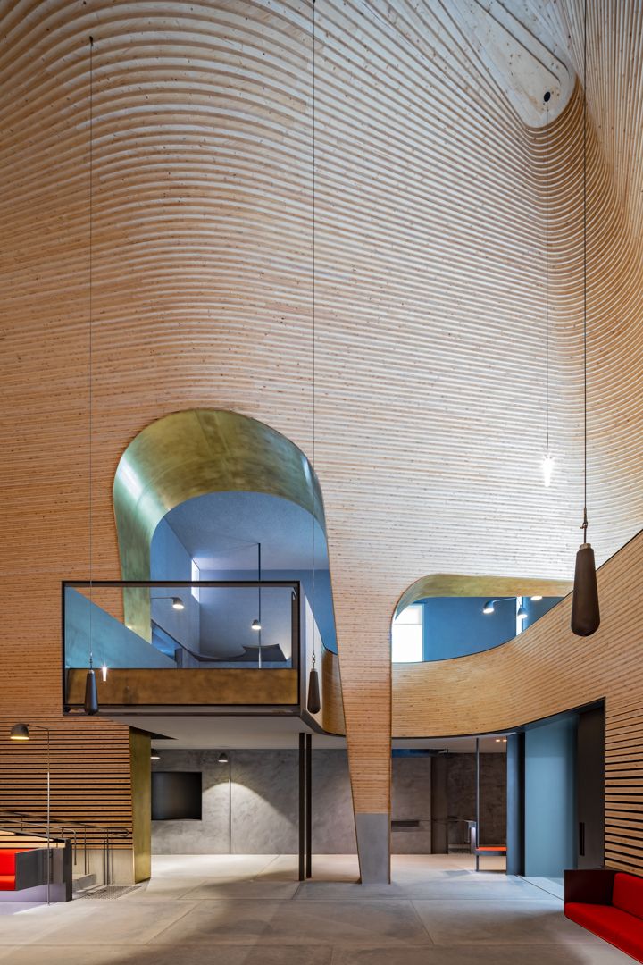 Dramatic timber-clad atrium with curved wooden walls, gold-lined archways, blue interior rooms, glass balustrade and pendant lights.
