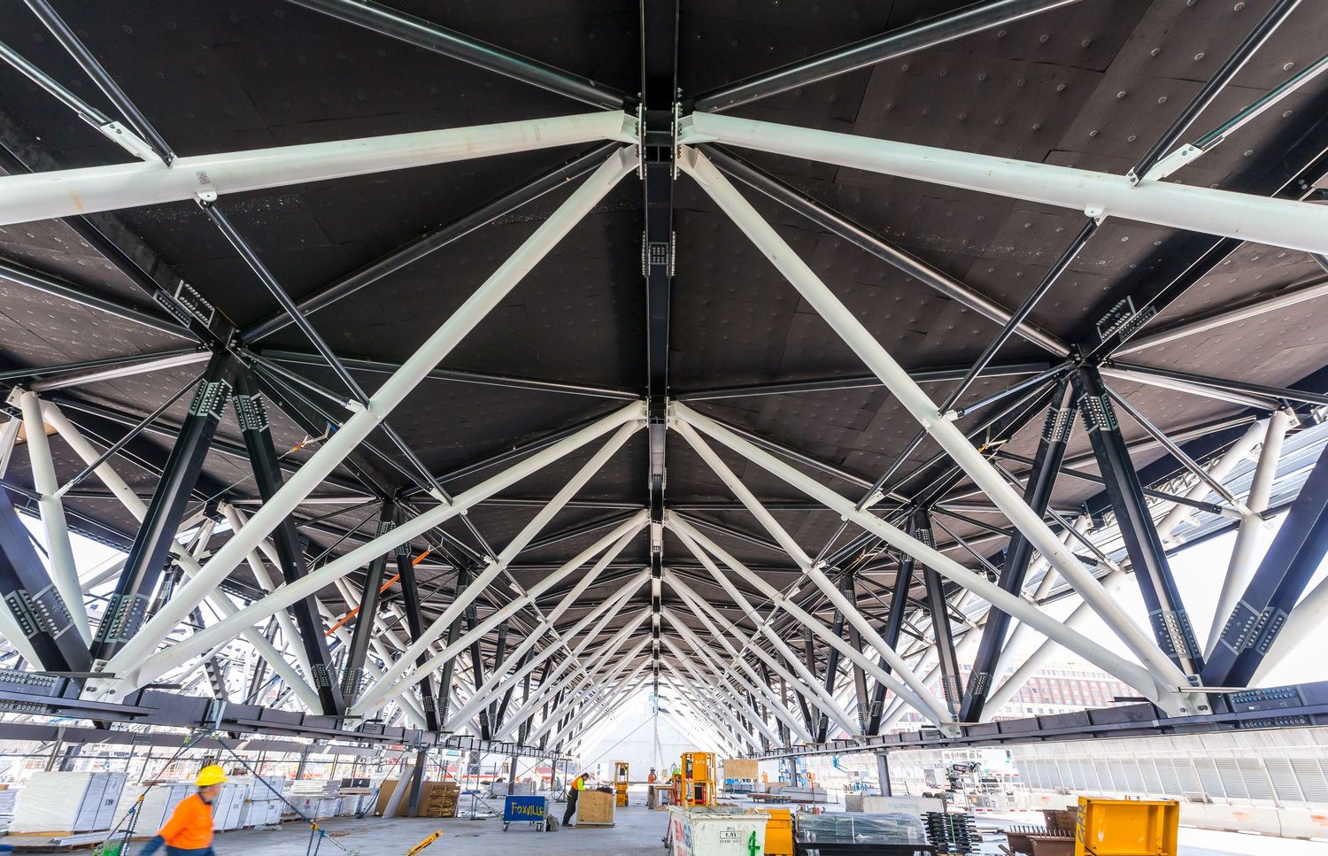 Large steel truss roof structure with white diagonal beams and dark cross bracing above a construction site.