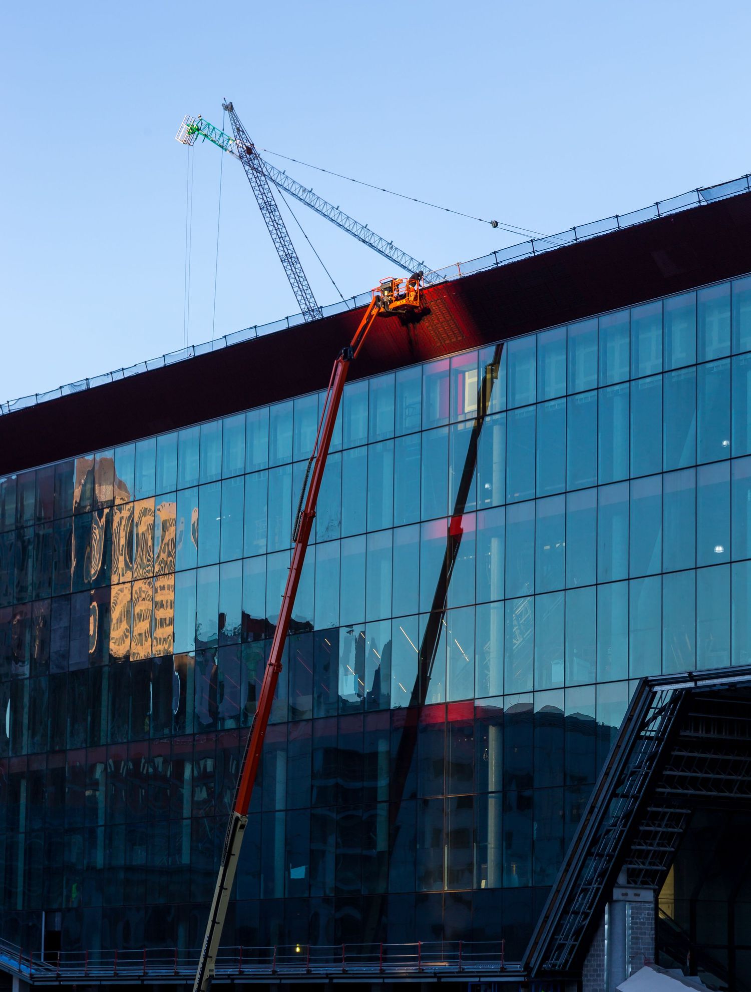 Red cherry picker and tower crane beside a glass-fronted building under construction, reflecting warm light at dusk.