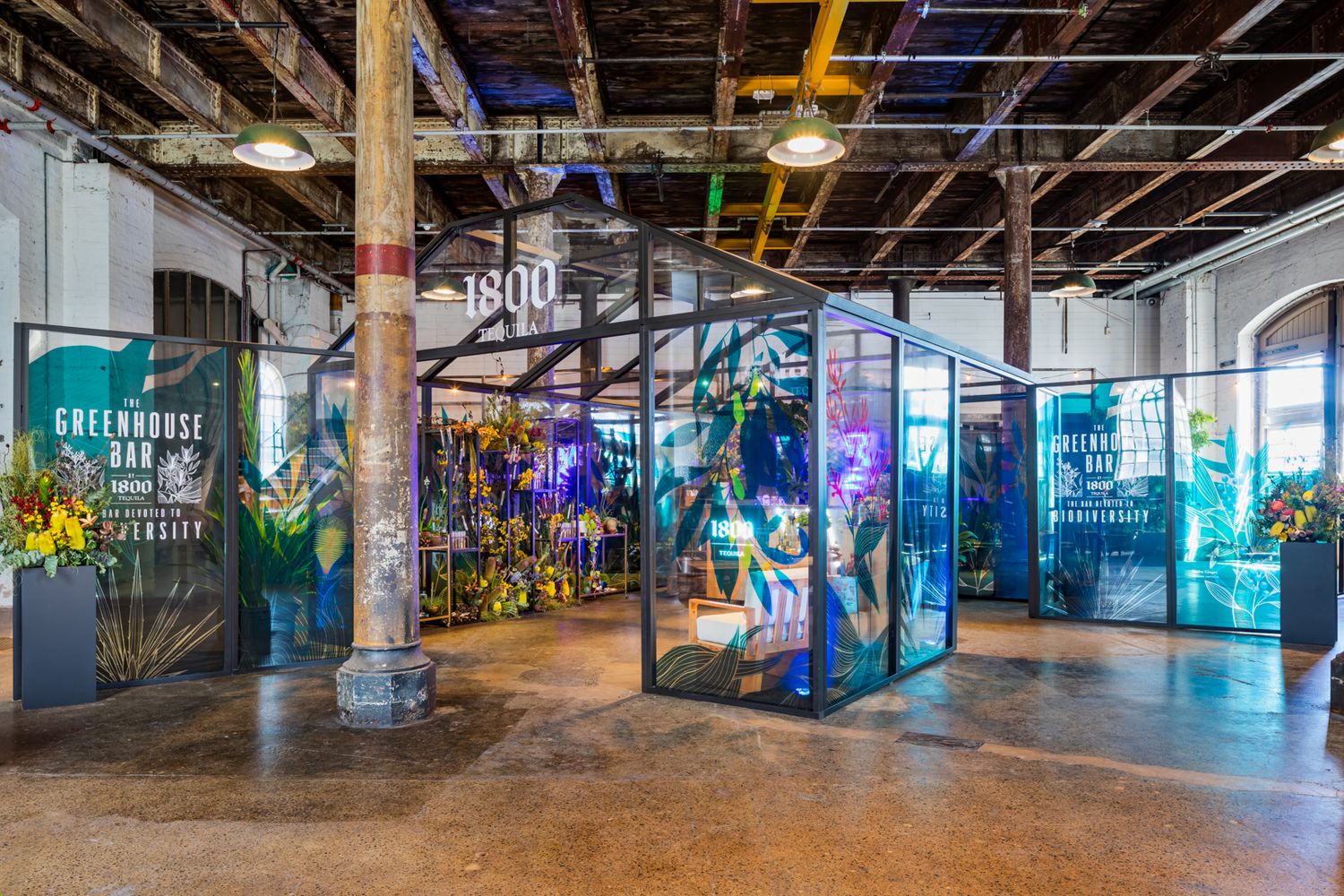 Industrial warehouse event space with glass greenhouse display structures, floral arrangements and exposed timber beams.