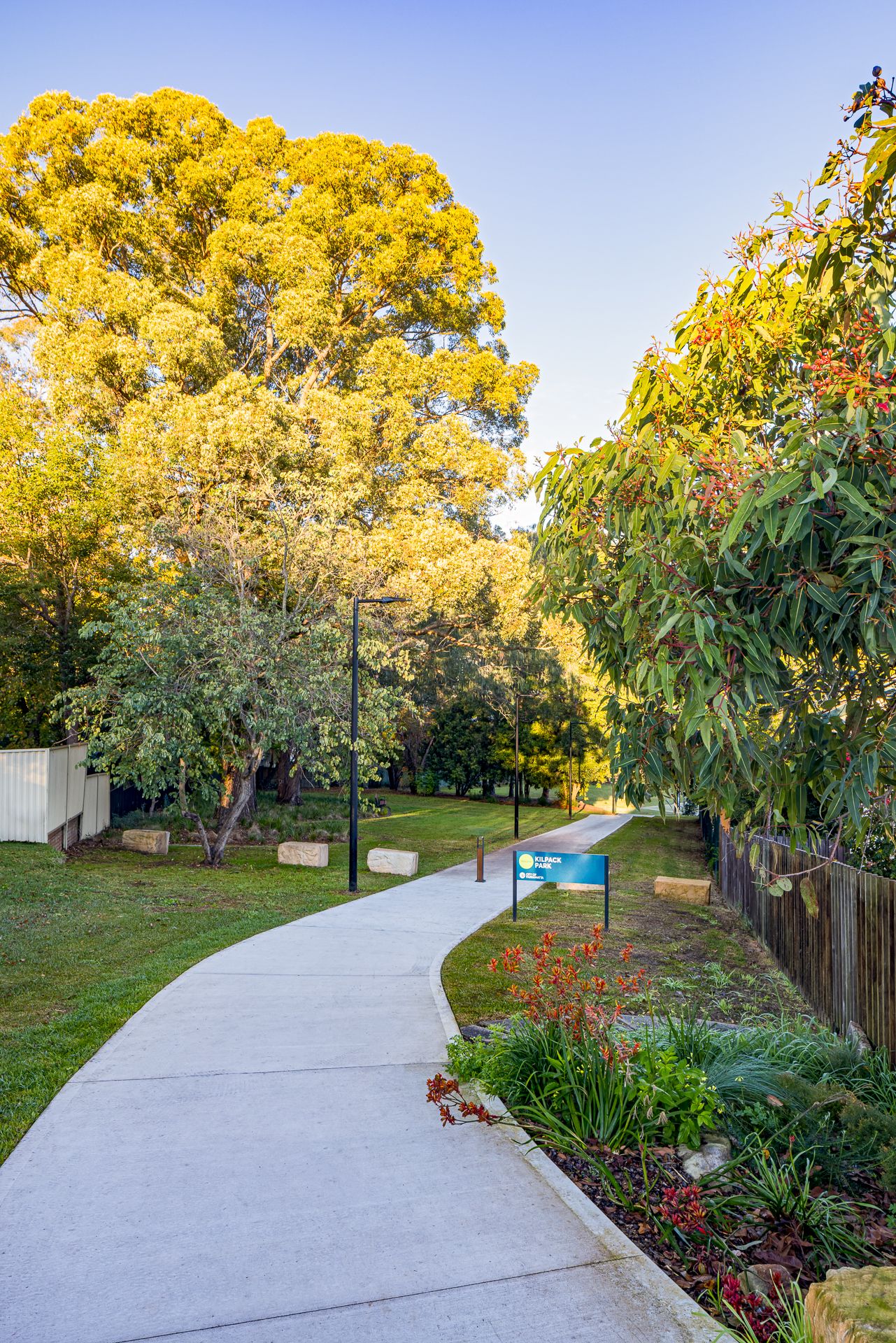 Curving concrete footpath through a sunlit park lined with colorful native plants, gum trees, lamp posts and a blue park sign.