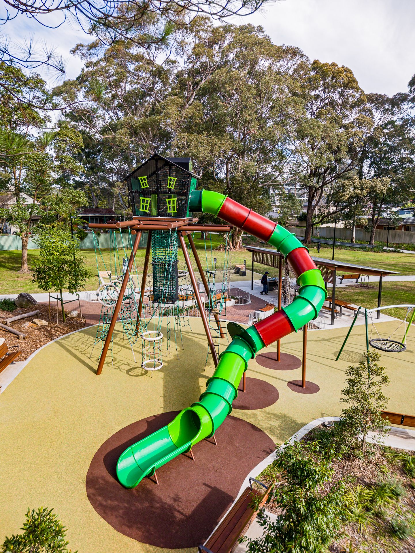 Colorful playground with a green and red tube slide, wooden tower, climbing nets and swings surrounded by eucalyptus trees.