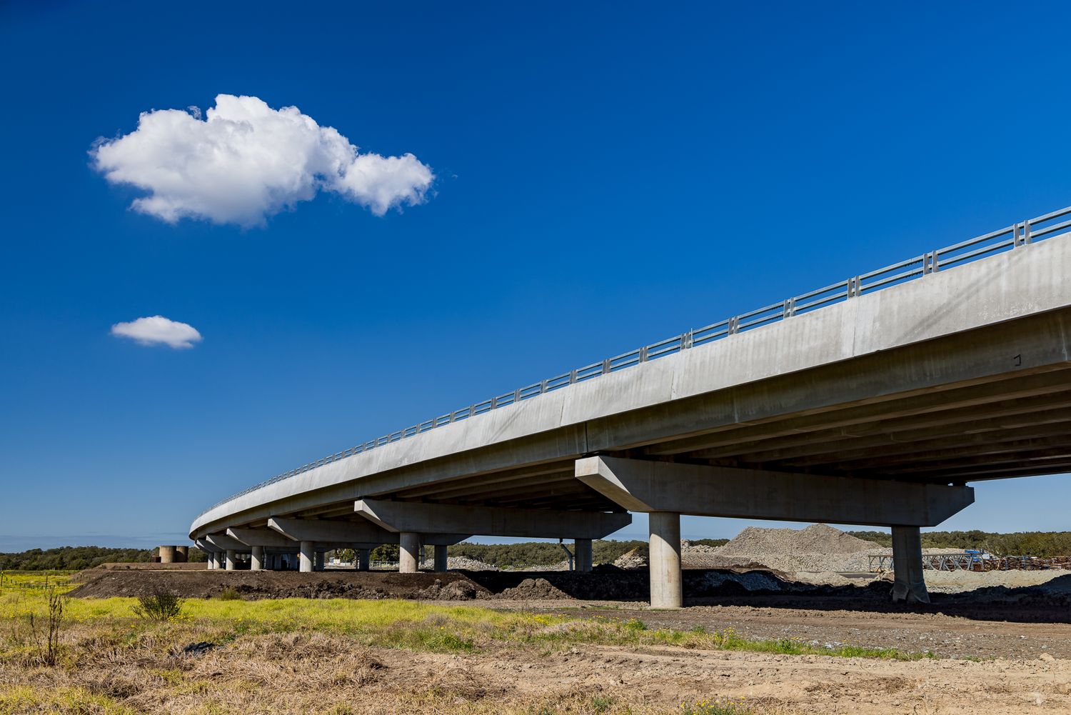 Curved concrete highway overpass supported by angled piers above dry grassland and dirt, with a lone white cloud in a blue sky.