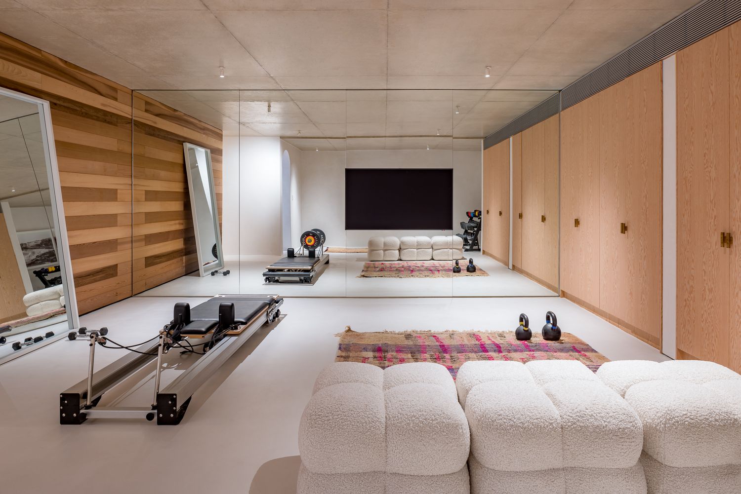 Spacious home gym and lounge with Pilates reformer, wood-paneled walls, bouclé sofa, wall mirrors and a large television.