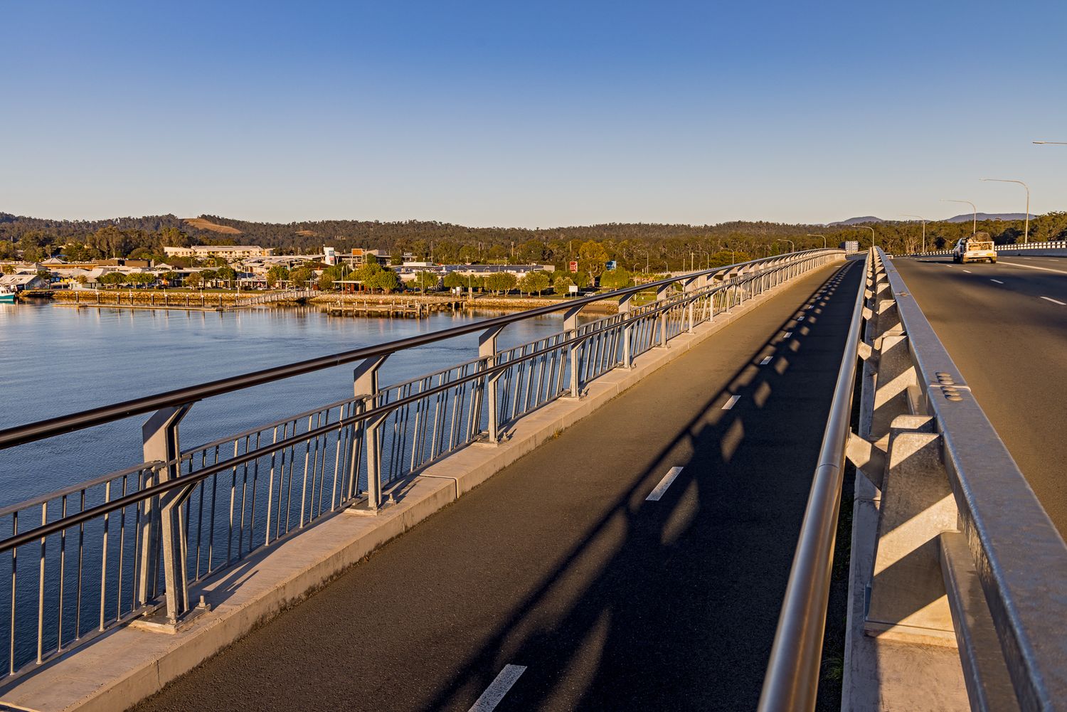 Pedestrian and cycle path alongside a bridge railing overlooking a calm bay and a coastal town on the shore in warm evening light.