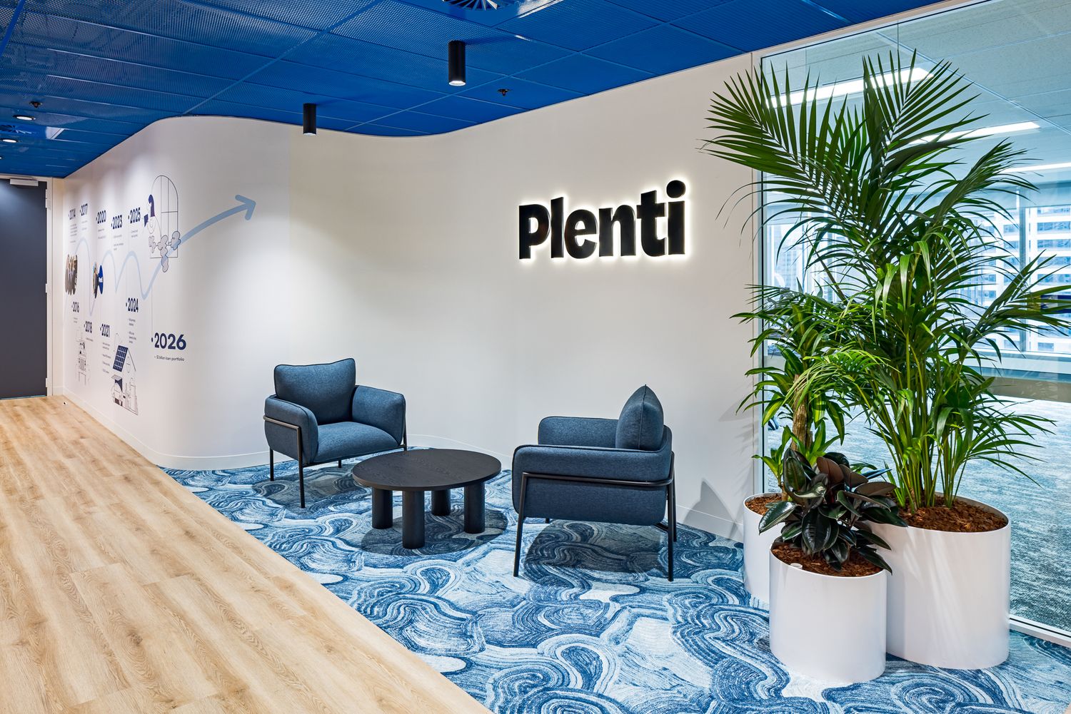 Plenti office reception lounge with two navy armchairs, a round coffee table, potted palms and an illuminated company logo.